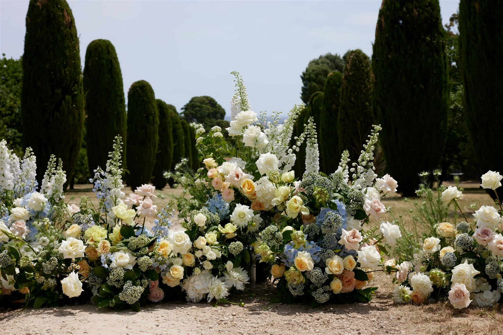 Large bouquet of white and pastel-colored flowers in front of a row of tall, dark green trees under a clear, partly cloudy sky.