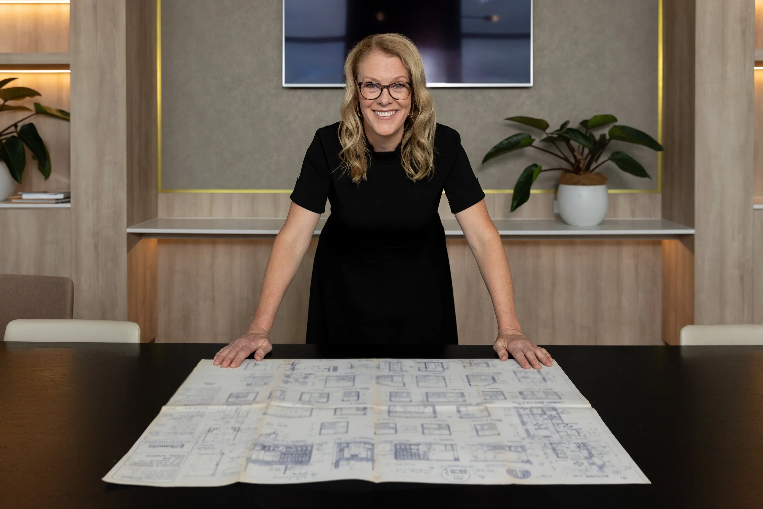 A woman with blonde hair, glasses, and a black shirt, smiling, leaning forward on a table with blueprints or architectural plans, in a modern office or conference room with potted plants and a mounted TV screen in the background.