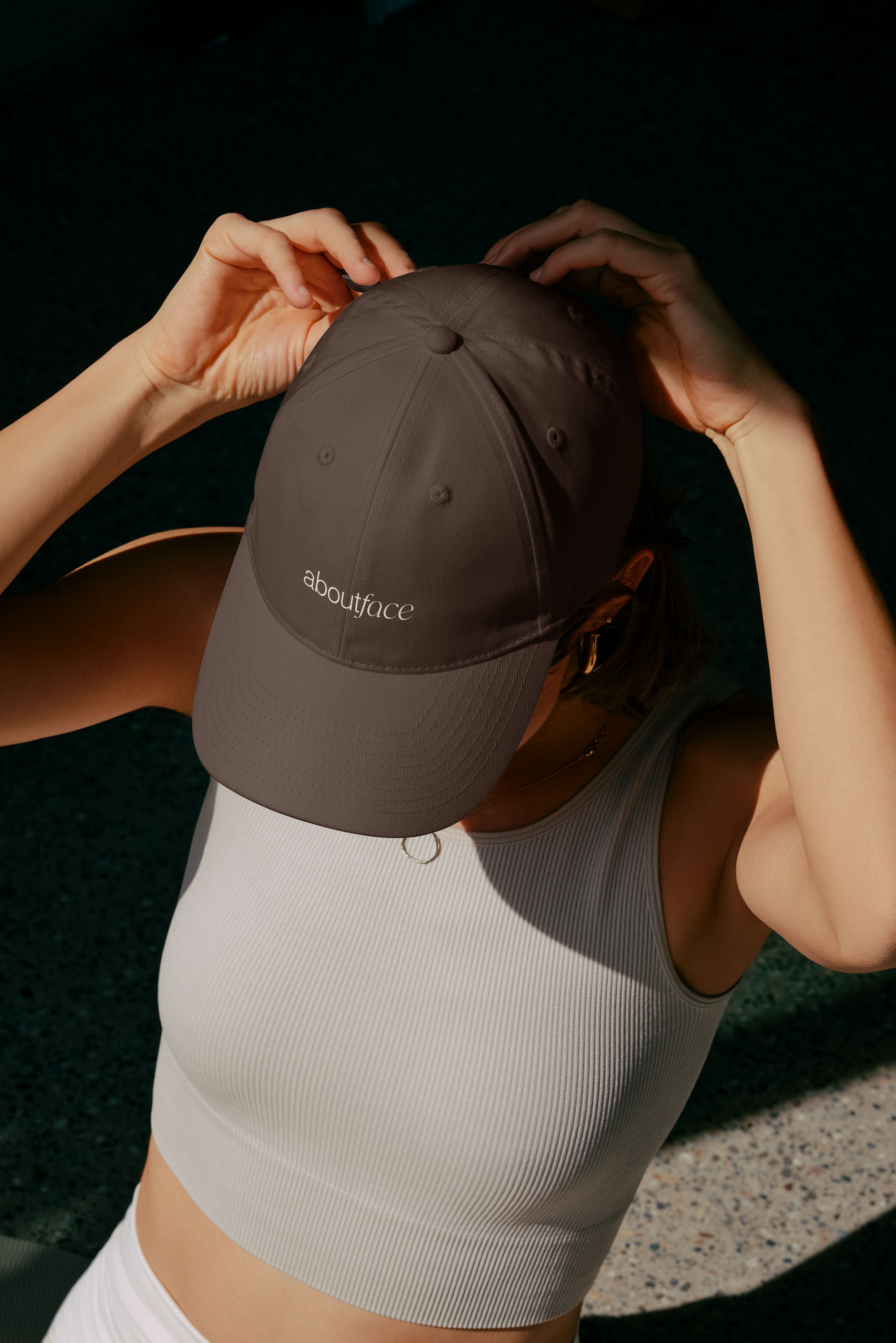 Person wearing a gray baseball cap with the words 'aboutface' on the front, white sleeveless top, and white pants, holding the brim of the cap with both hands, against a dark textured background.