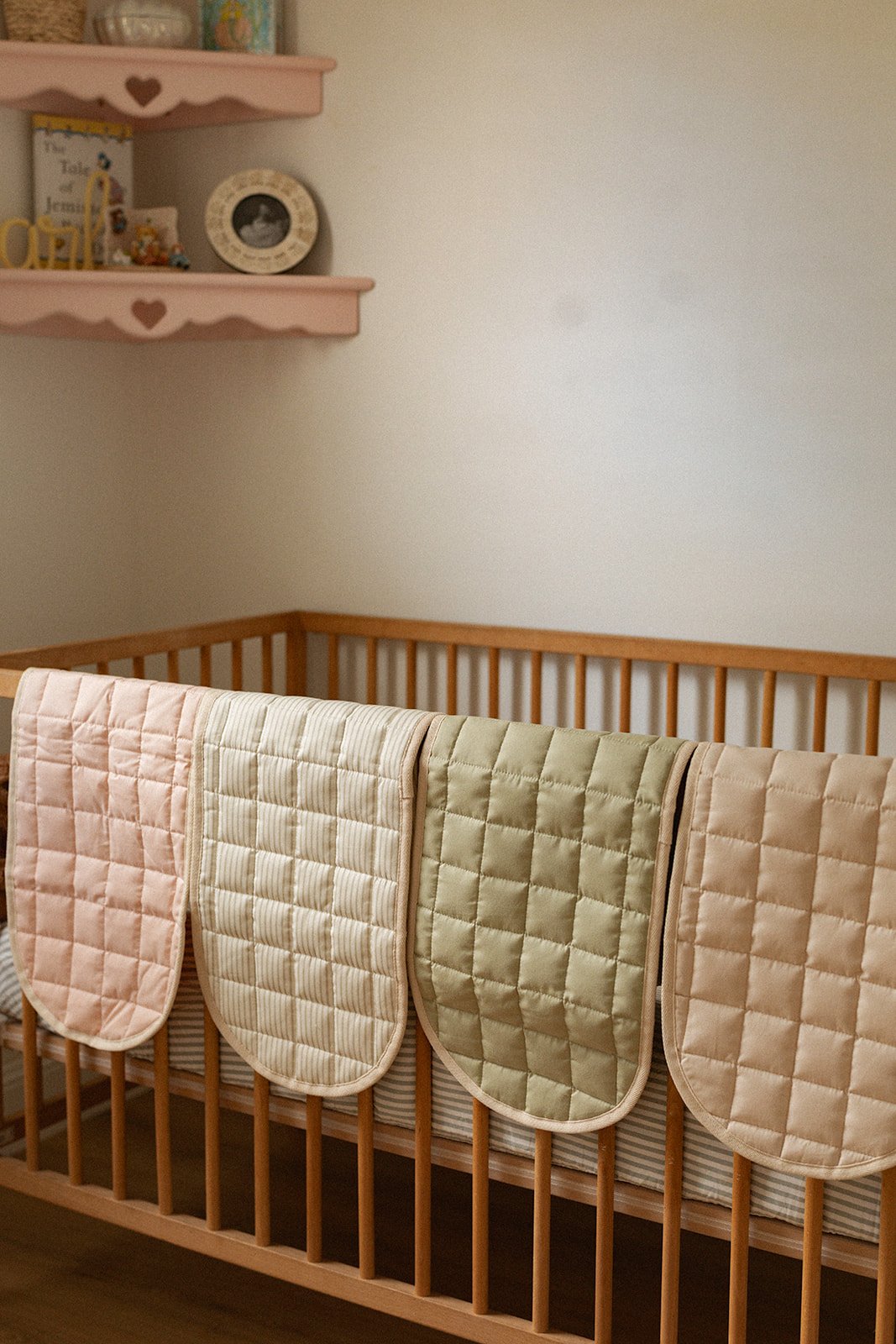 A wooden crib with four quilted mattress pads hanging over the side in pink, cream, light green, and peach colors, with a pink shelf above holding decorations and a framed photograph.