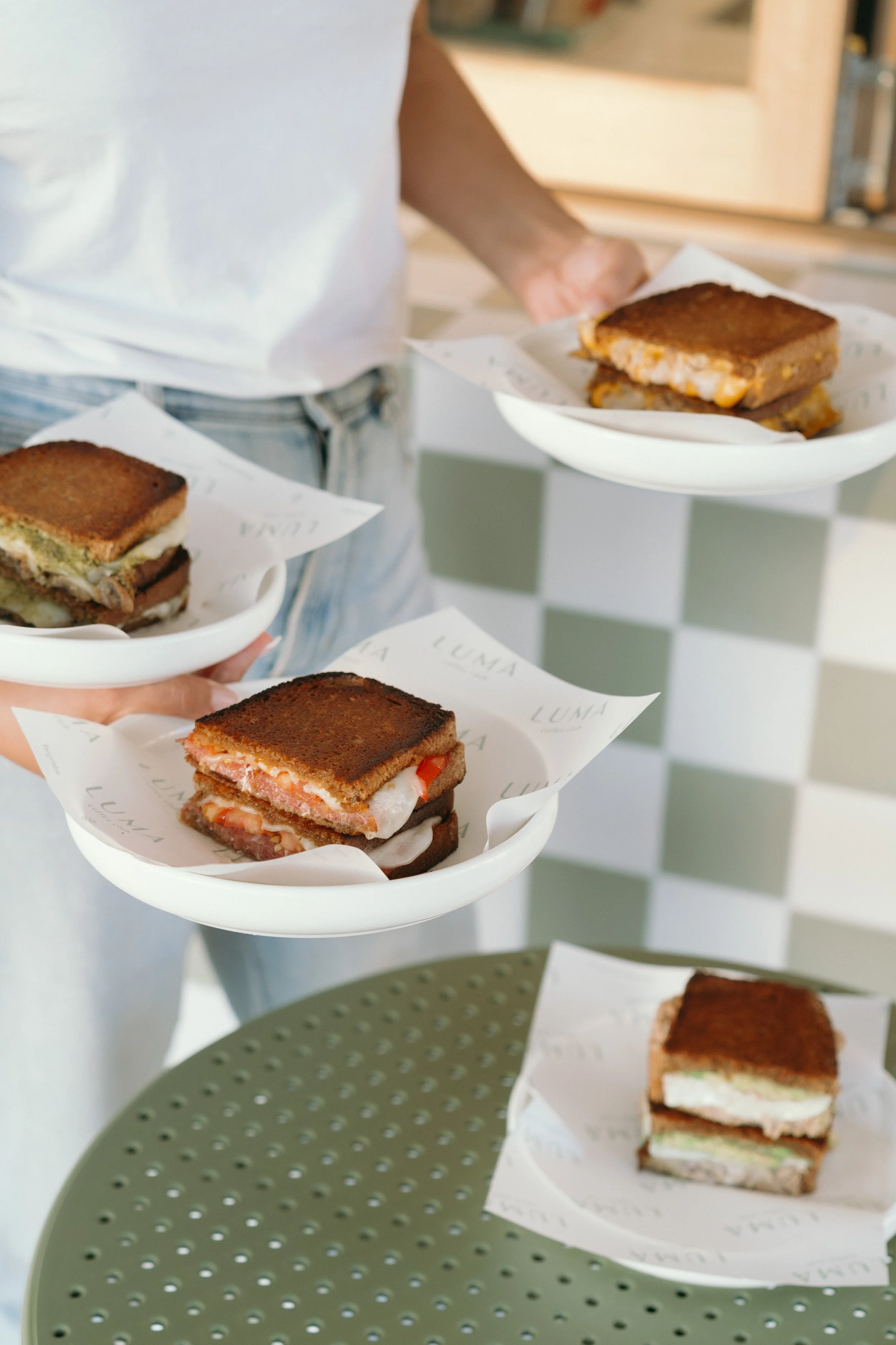 Multiple sandwiches with different fillings on white plates, served on parchment paper, held by a person wearing a white shirt. A checkered wall is in the background.