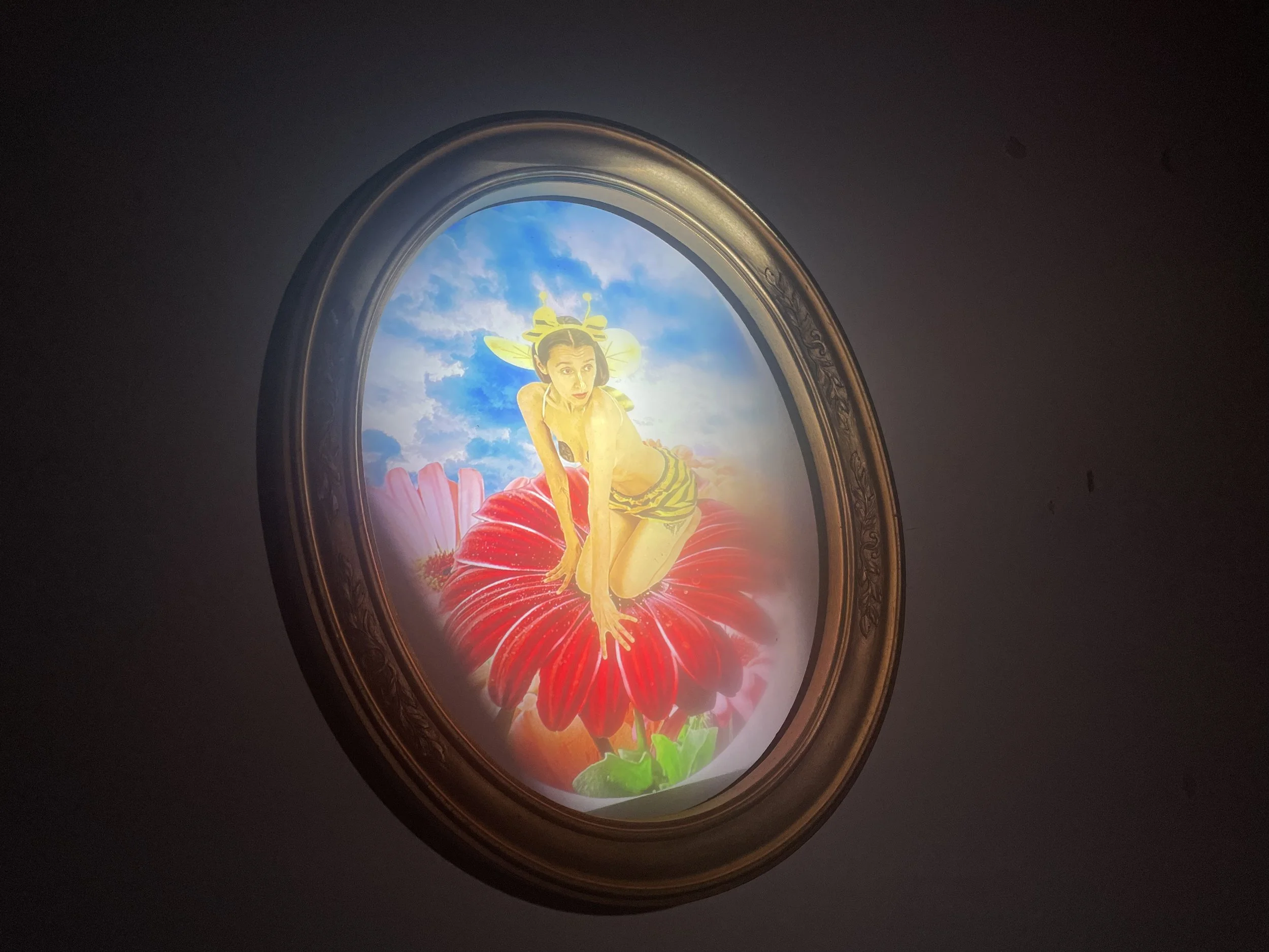 A photograph of a framed composited image. The image features a yellow tinted woman sitting on a flower, appearing similar to a bumble bee. The colors of the portrait are very bright and reminiscent of a children's book illustration.