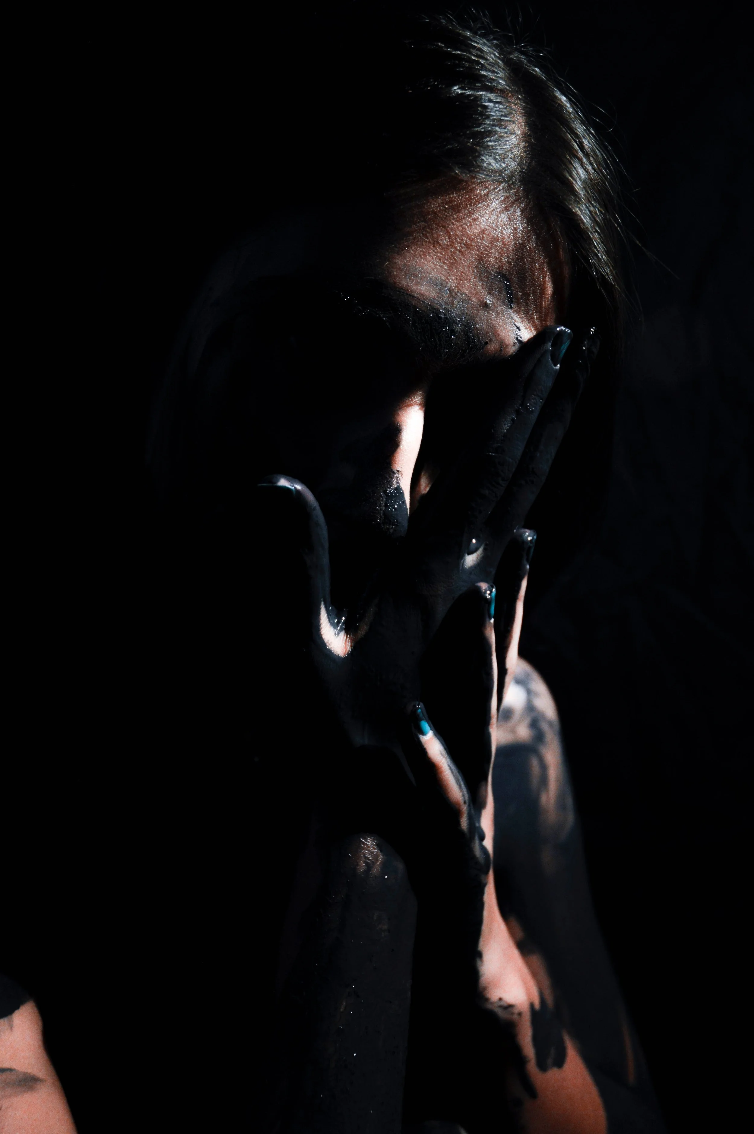 A dramatic photograph of a person holding their paint covered hands to their face. The bright but minimal lighting reveals only small portions of the person's face, the light bouncing off their hair, forehead, nose, and the paint on their fingertips