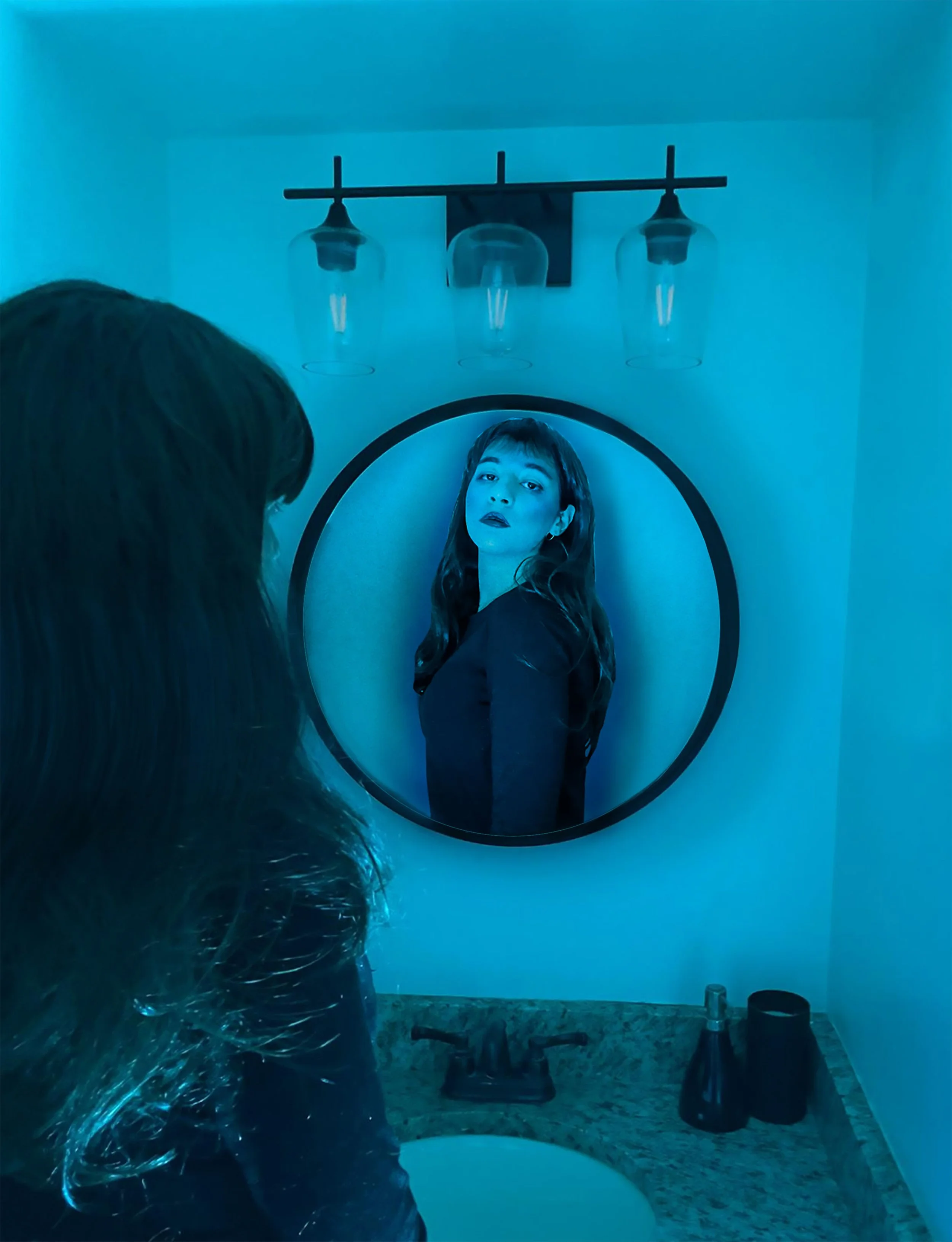 A photo composite of a girl standing in front of a bathroom sink, looking at her reflection in a mirror.  The room lighting is blue, and the girl's pose in the foreground is different than the one in the reflection.