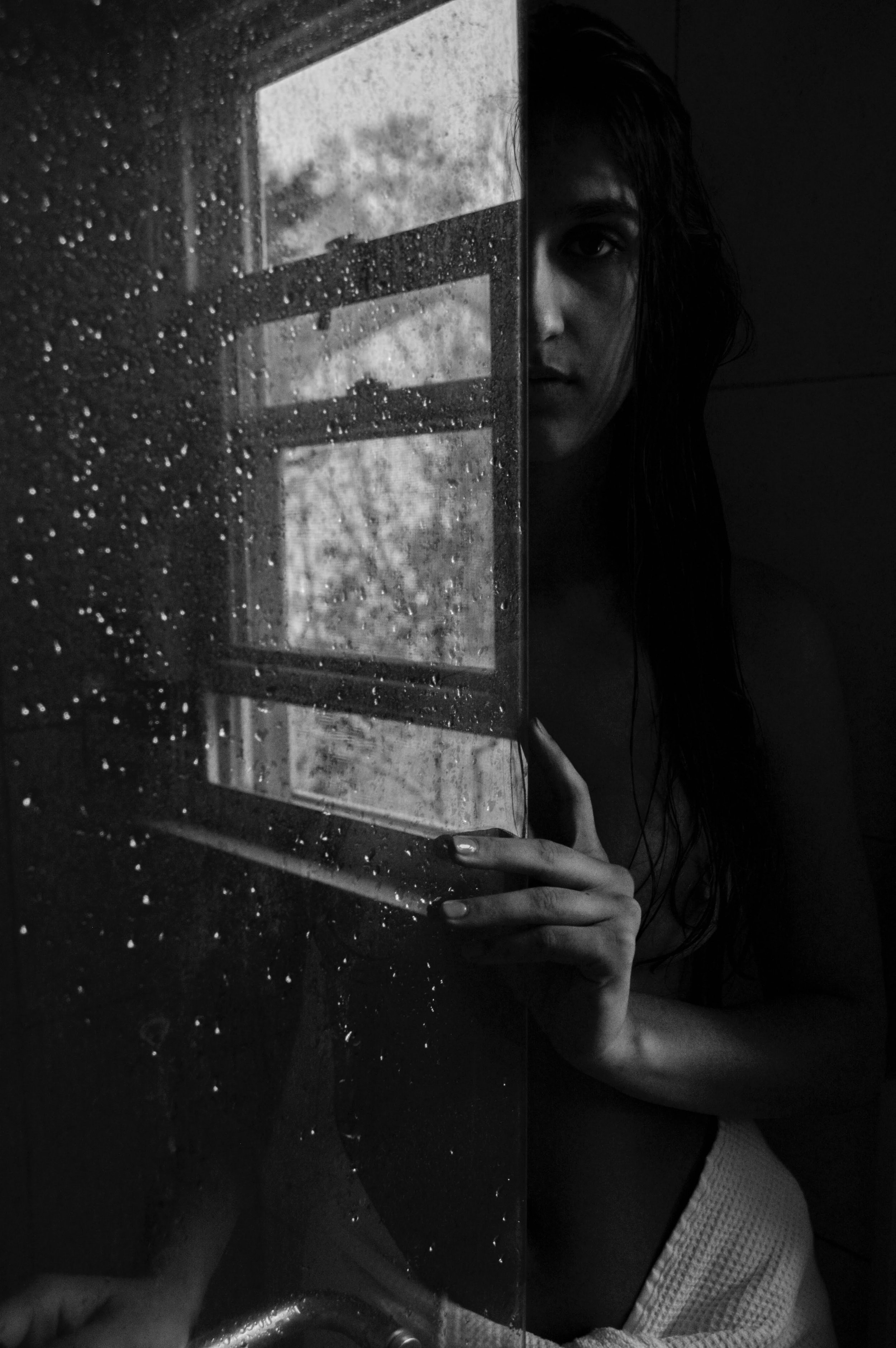 A black and white digital photograph of a girl peaking out from behind a glass door. The girl is standing in shadow, and the glass door covered in water droplets reflects an open window to the outside. Her fingers rest gently on the glass.