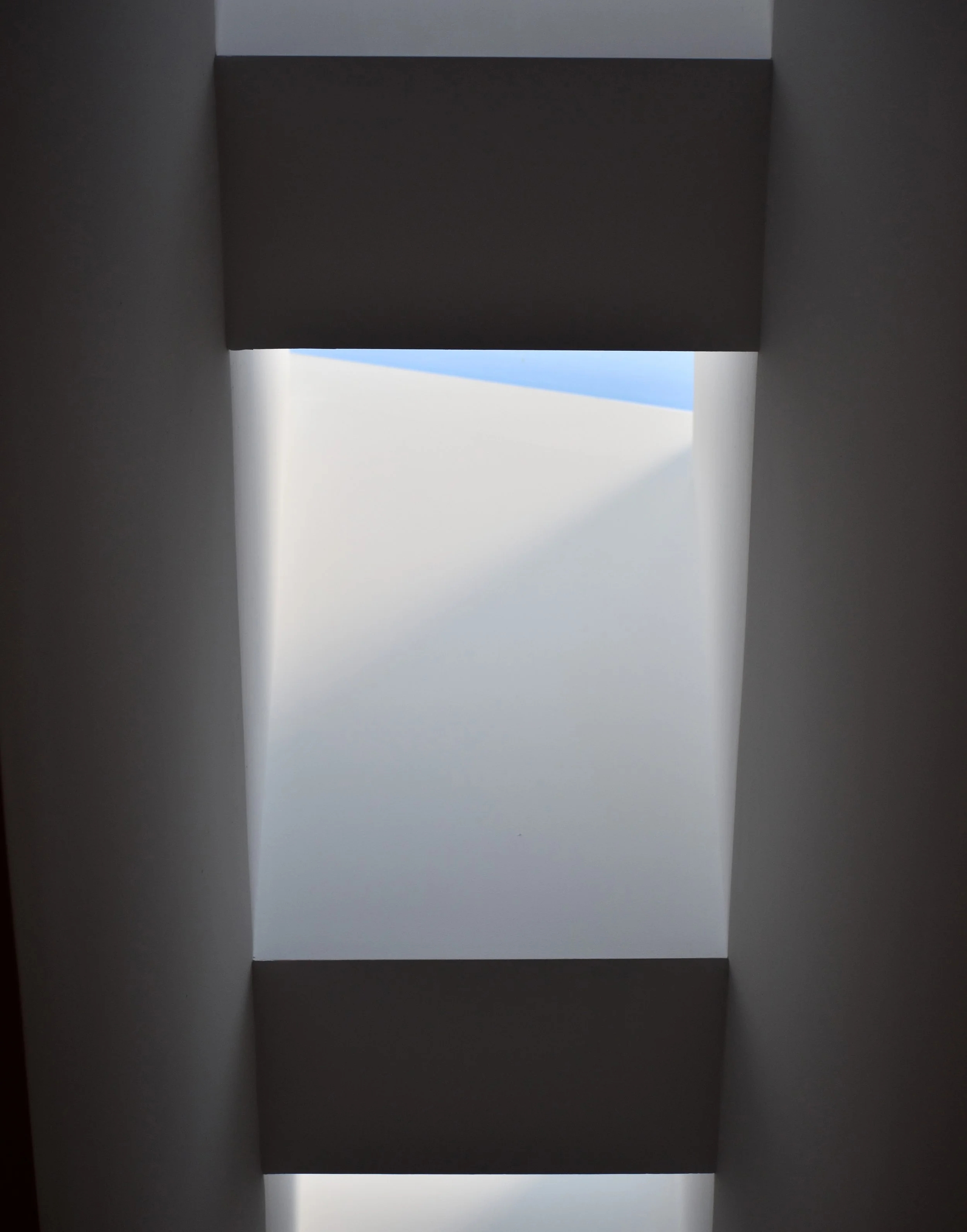 A photo of a rectangular gap in the ceiling, as seen from below. The architecture is white and there is a triangular sliver of blue sky peeking through the gap. light comes in at a neat angle, making the piece appear very geometric and clean.