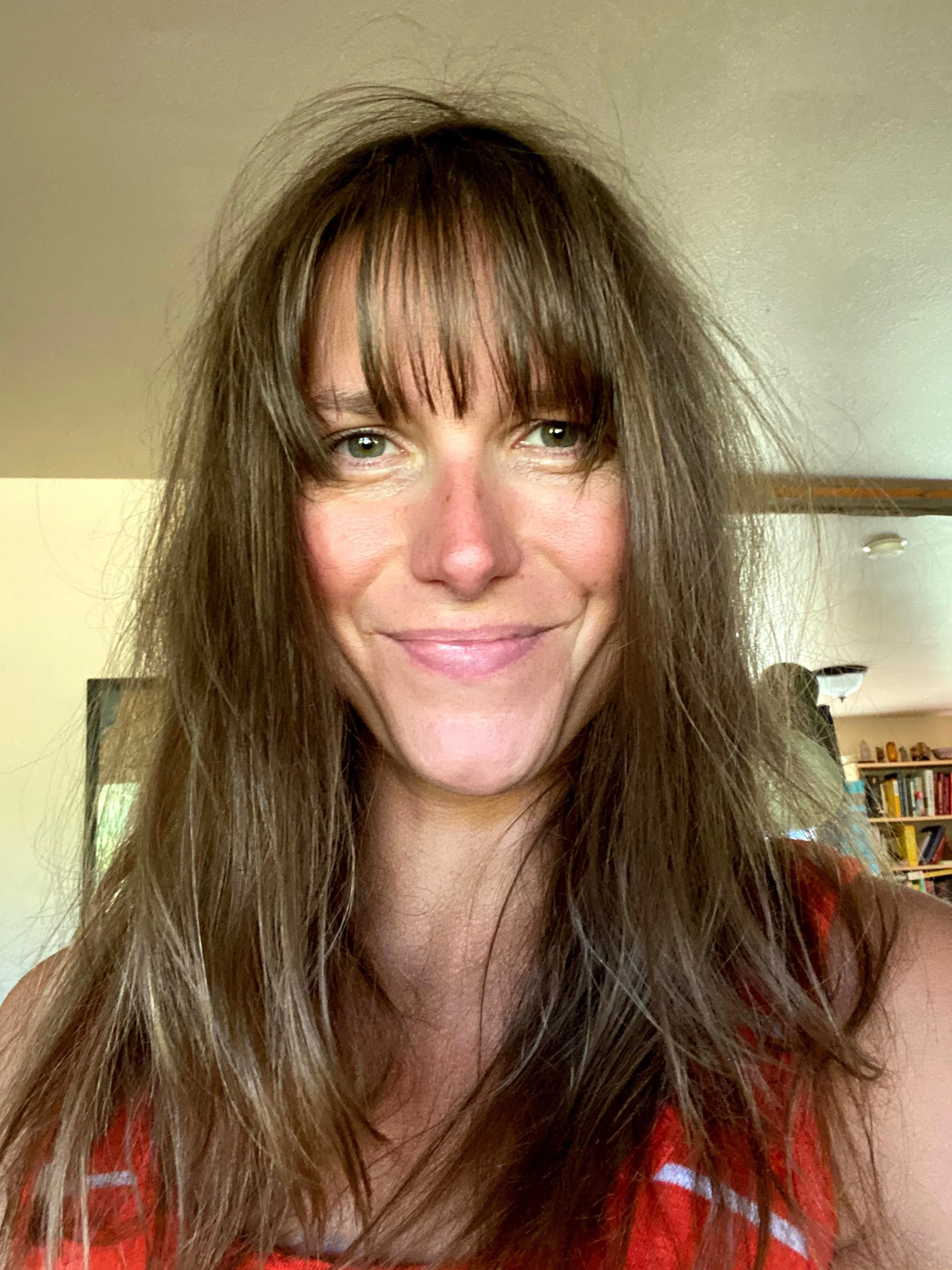 Close-up of a woman with long, wavy brown hair and bangs, smiling. She has fair skin and light green eyes. She is wearing a red top with white patterns, indoors with bookshelves in the background.