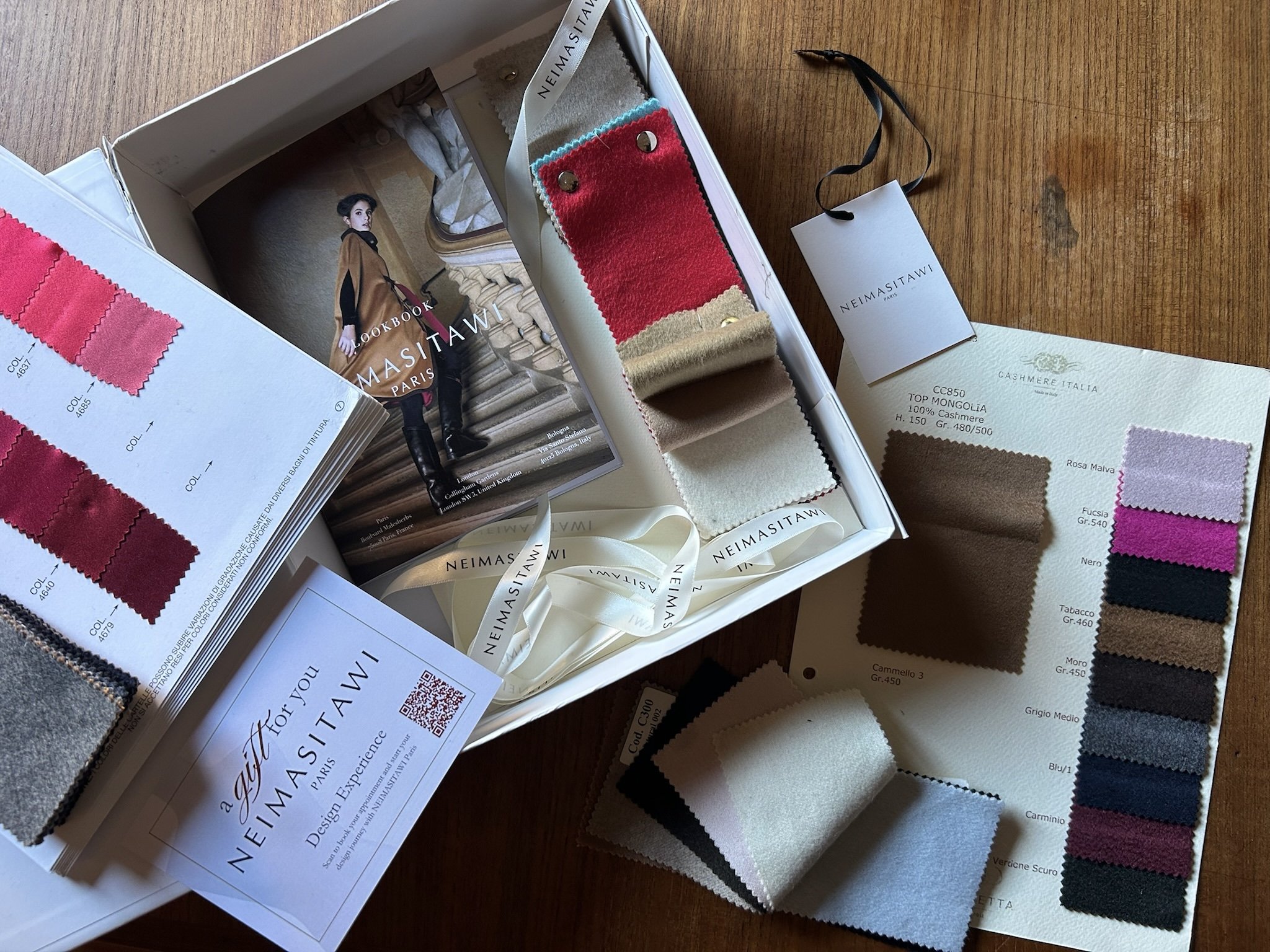 A collection of fabric swatches, color sample cards, and a brochure from Neimasitawi on a wooden surface, showcasing various fabric colors and textures for potential clothing or interior design.