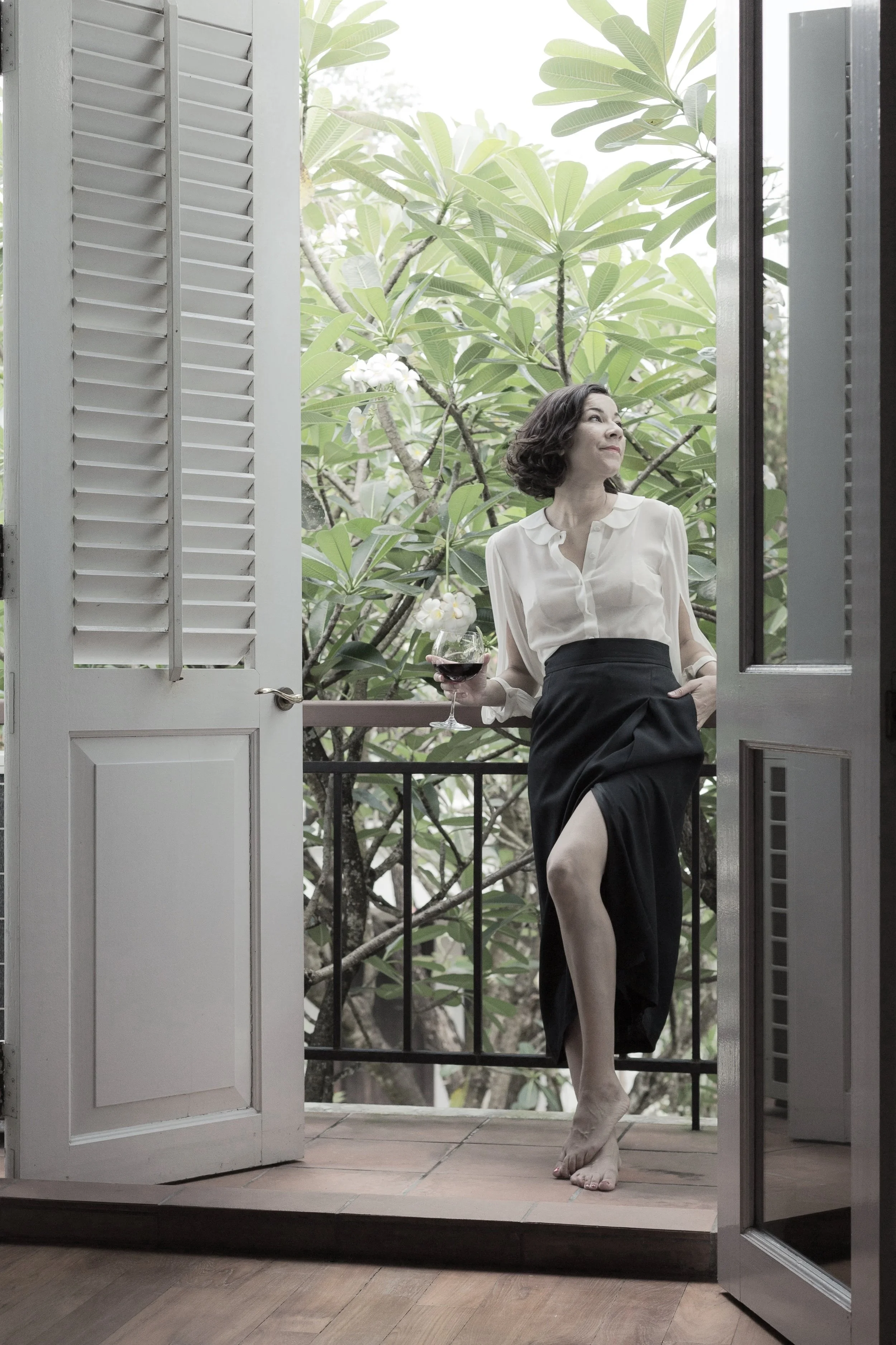 Woman standing on a balcony with a glass of red wine, wearing a white blouse and black skirt, looking to the side with greenery in the background.