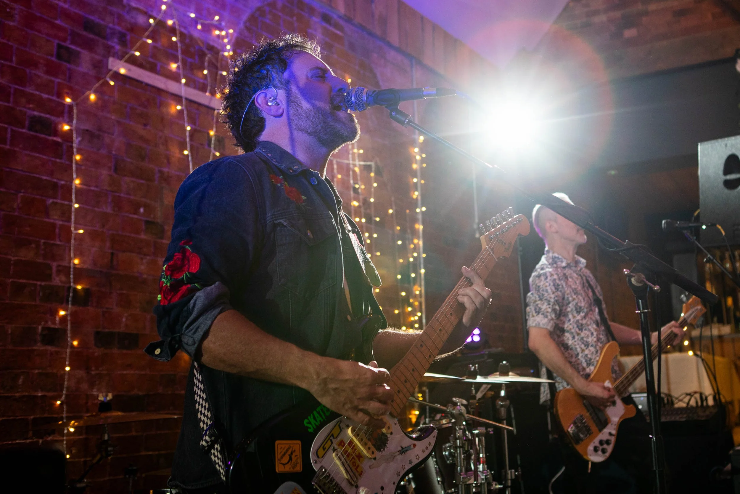 Two musicians performing on stage with brick wall backdrop and string lights. One playing guitar and singing into a microphone, the other playing bass guitar.