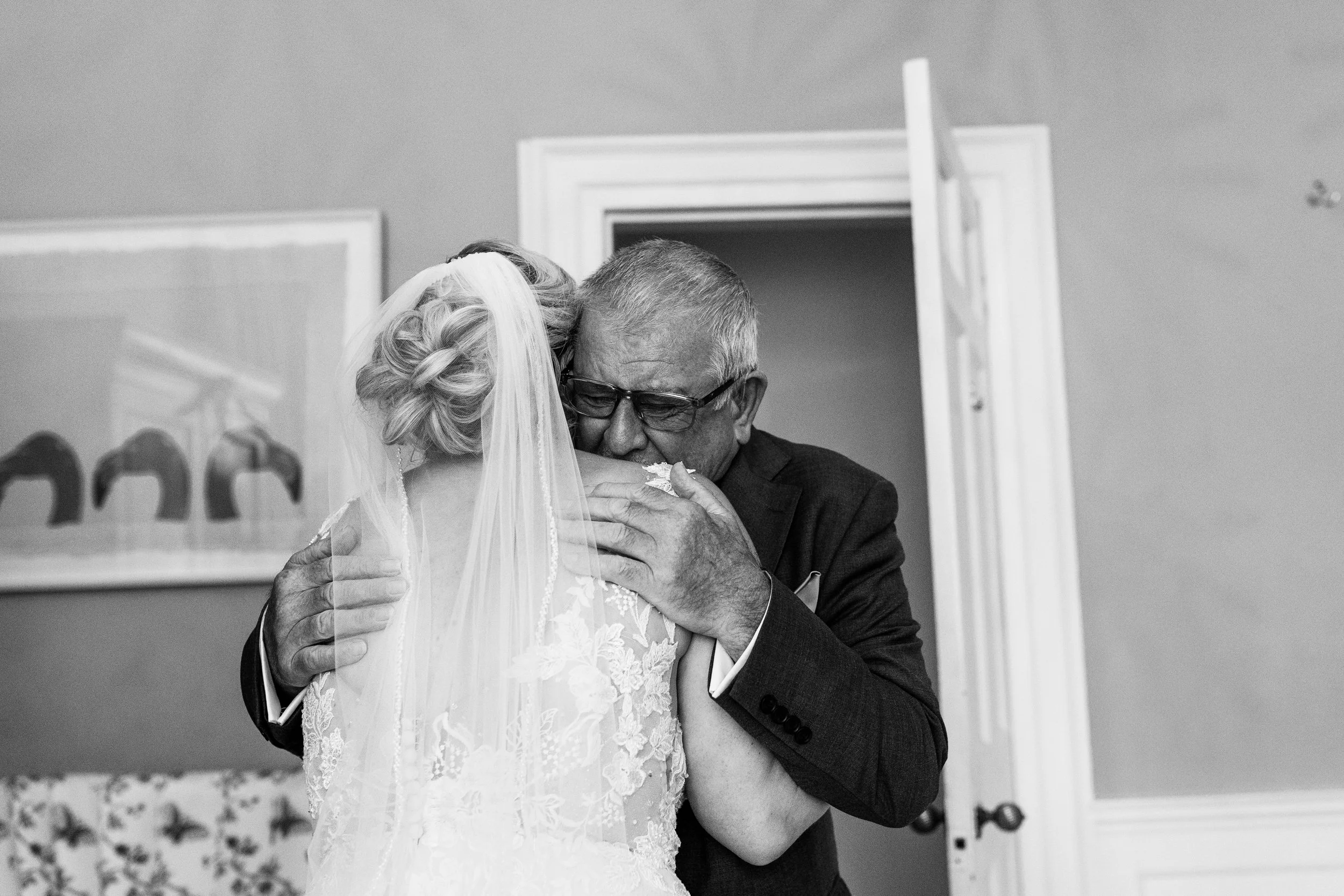 An emotional moment where a father, is hugging a bride, in a wedding dress with a veil. The scene is in a room with a doorway and two paintings on the wall.