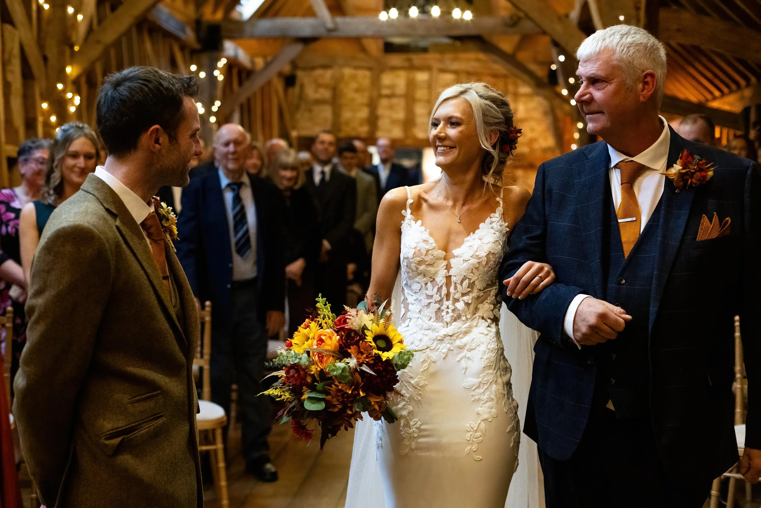 A bride in a white wedding gown holding a bouquet of yellow and red flowers walking down the aisle with her father in a dark suit, while the groom in a brown suit waits for her at the altar during a wedding ceremony in a rustic barn decorated with st