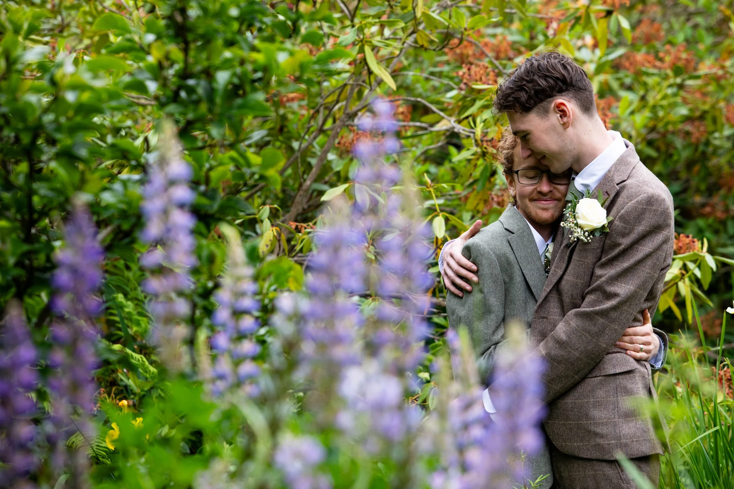 Two newly married grooms dressed in suits embracing outdoors in a lush garden with purple flowers in the foreground.