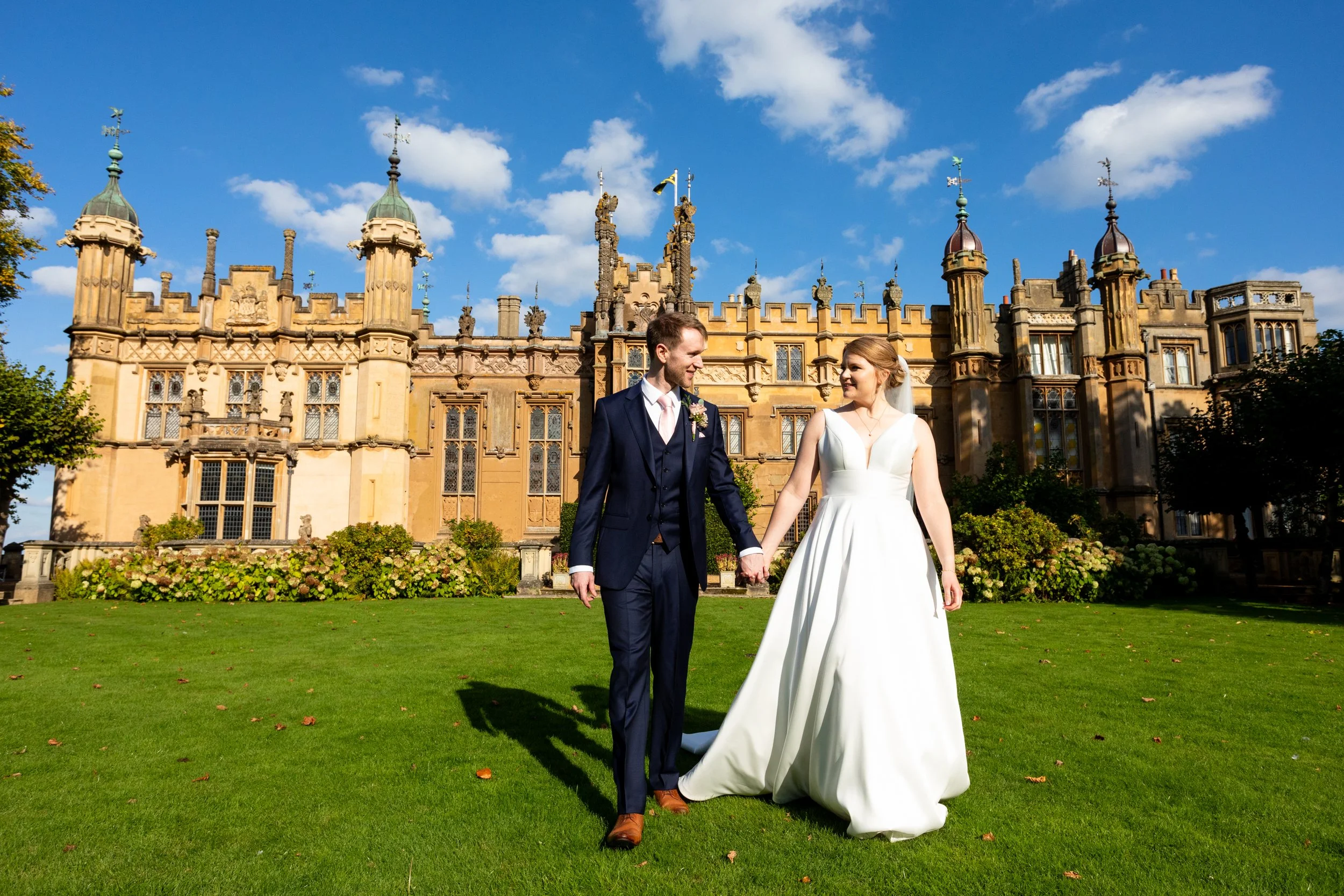 Newlywed couple holding hands and walking in front of the historic Knebworth House, a beautiful and iconic wedding venue in Hertfordshire. 