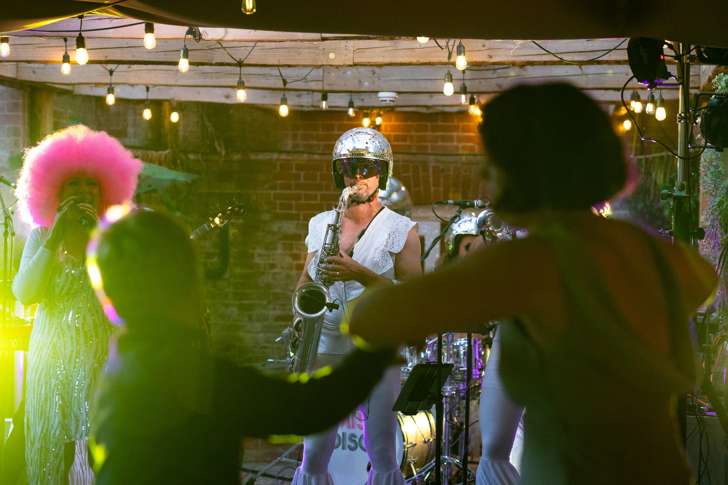 A band performing on stage with string lights hanging above. One woman has bright pink afro hair, wearing a sparkly striped dress, singing into a microphone. A man in the center is playing the saxophone, wearing a white outfit, a helmet, and sunglass