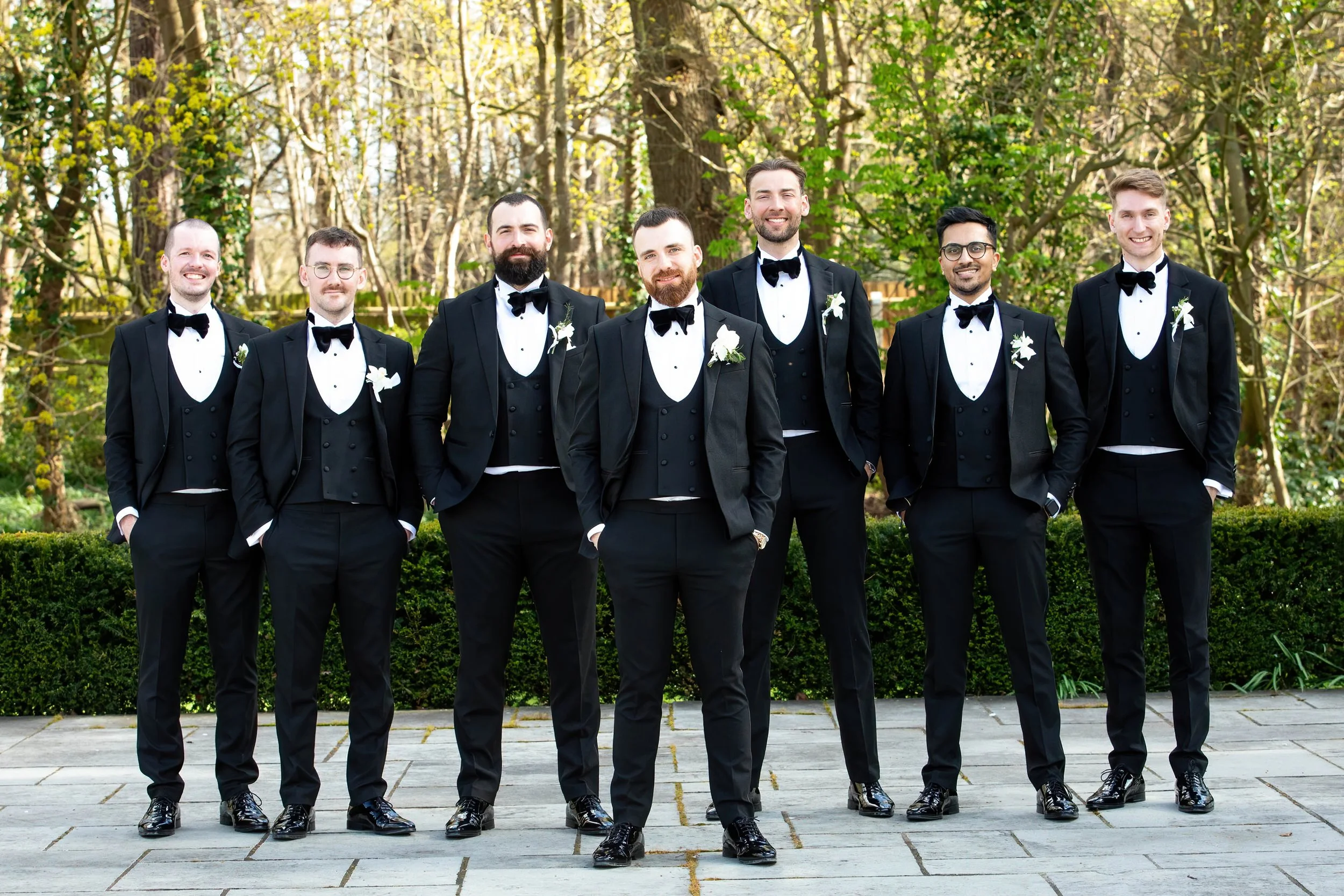 A group of eight men dressed in black tuxedos with bow ties, standing outdoors on a paved area with greenery and trees in the background.