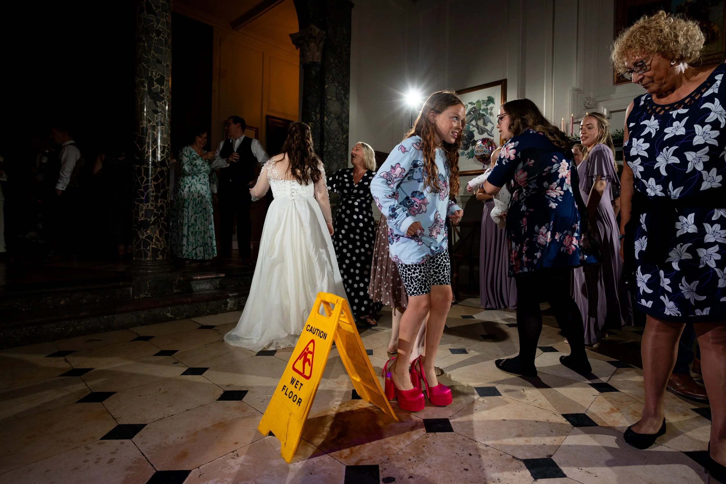 People dancing at a celebration, some wearing pajamas and others in formal attire. A woman in a white dress is in the background, and a woman in a blue floral dress is on the right. There is a yellow caution wet floor sign on the tiled floor.