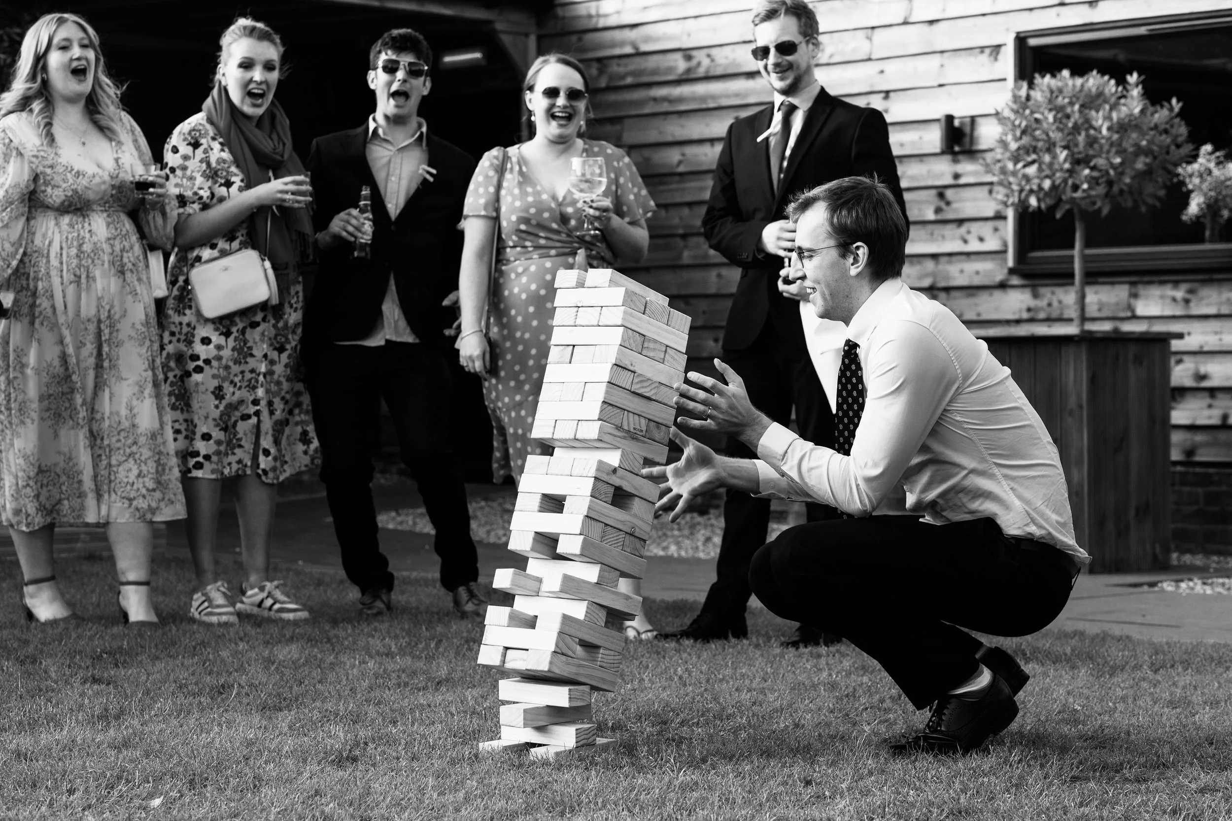 A man crouching and playing giant outdoor Jenga with guests watching and laughing at a garden party or social gathering.