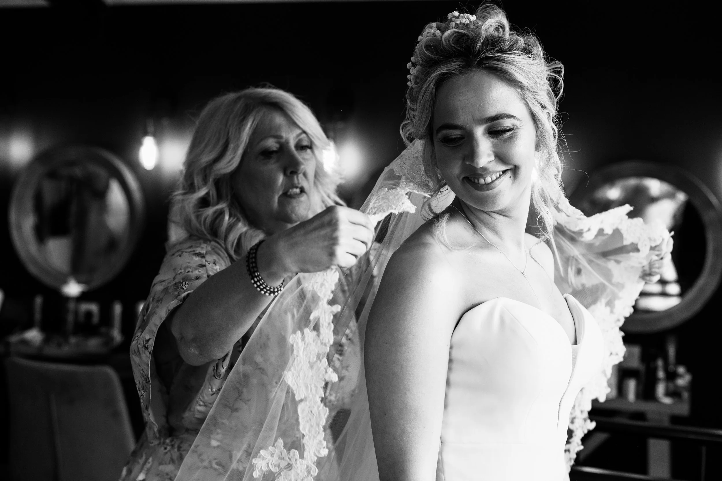 A bride smiling while her mother helps her put on her wedding veil in a dressing room.