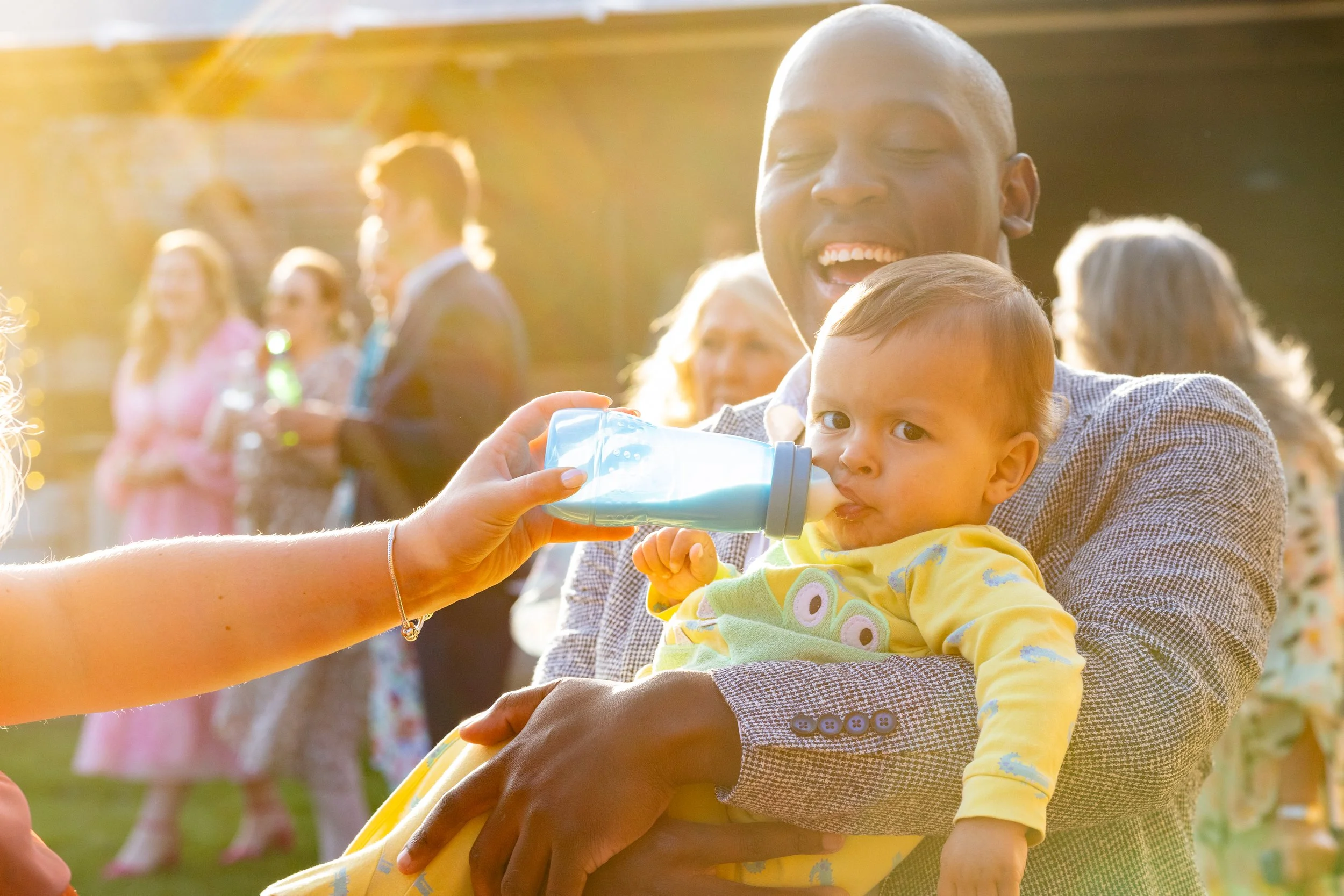 A man holding a baby while a woman feeds the baby with a bottle at an outdoor gathering in sunlight with people in the background.