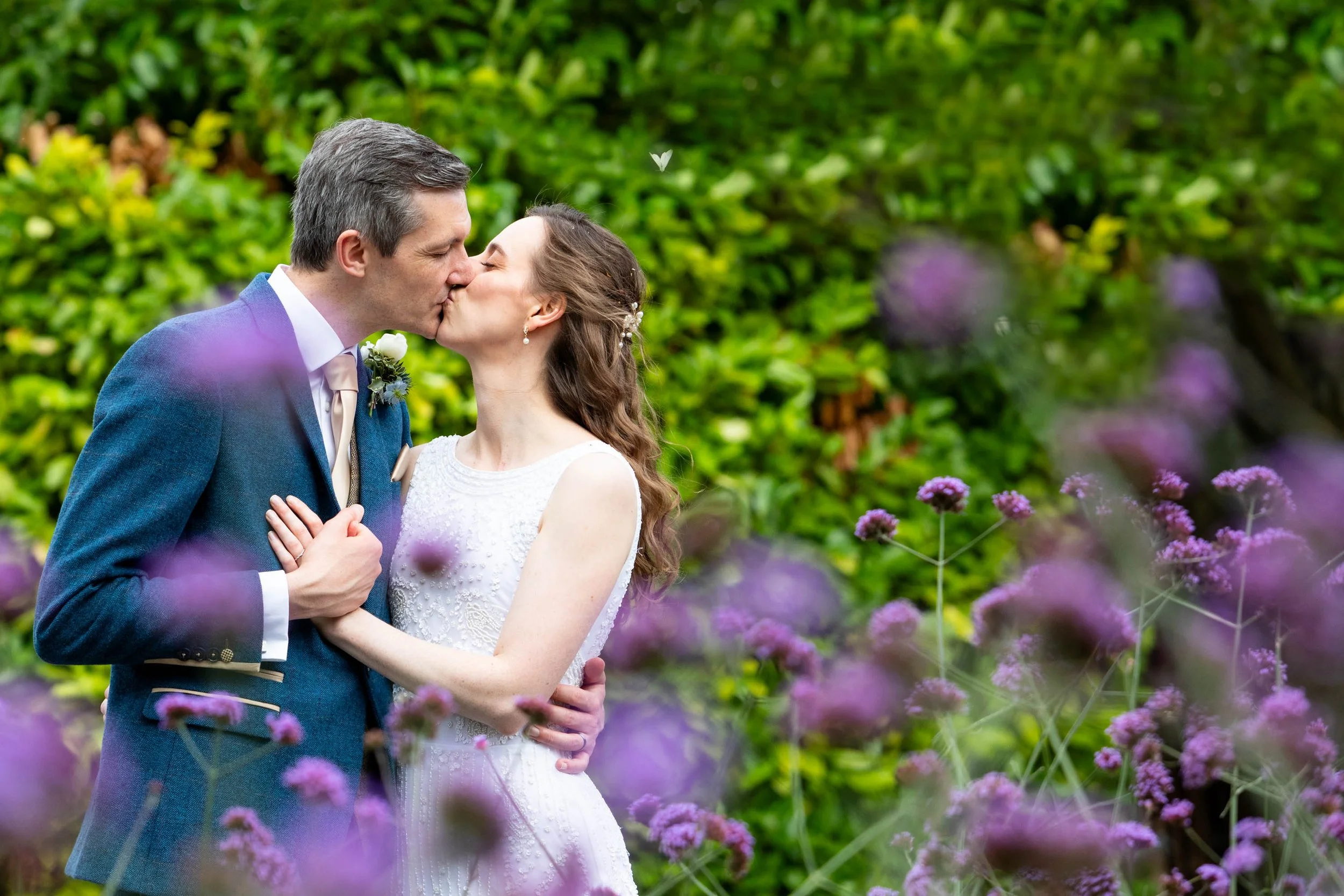 A newlywed couple sharing a kiss outdoors, surrounded by green bushes and purple flowers.