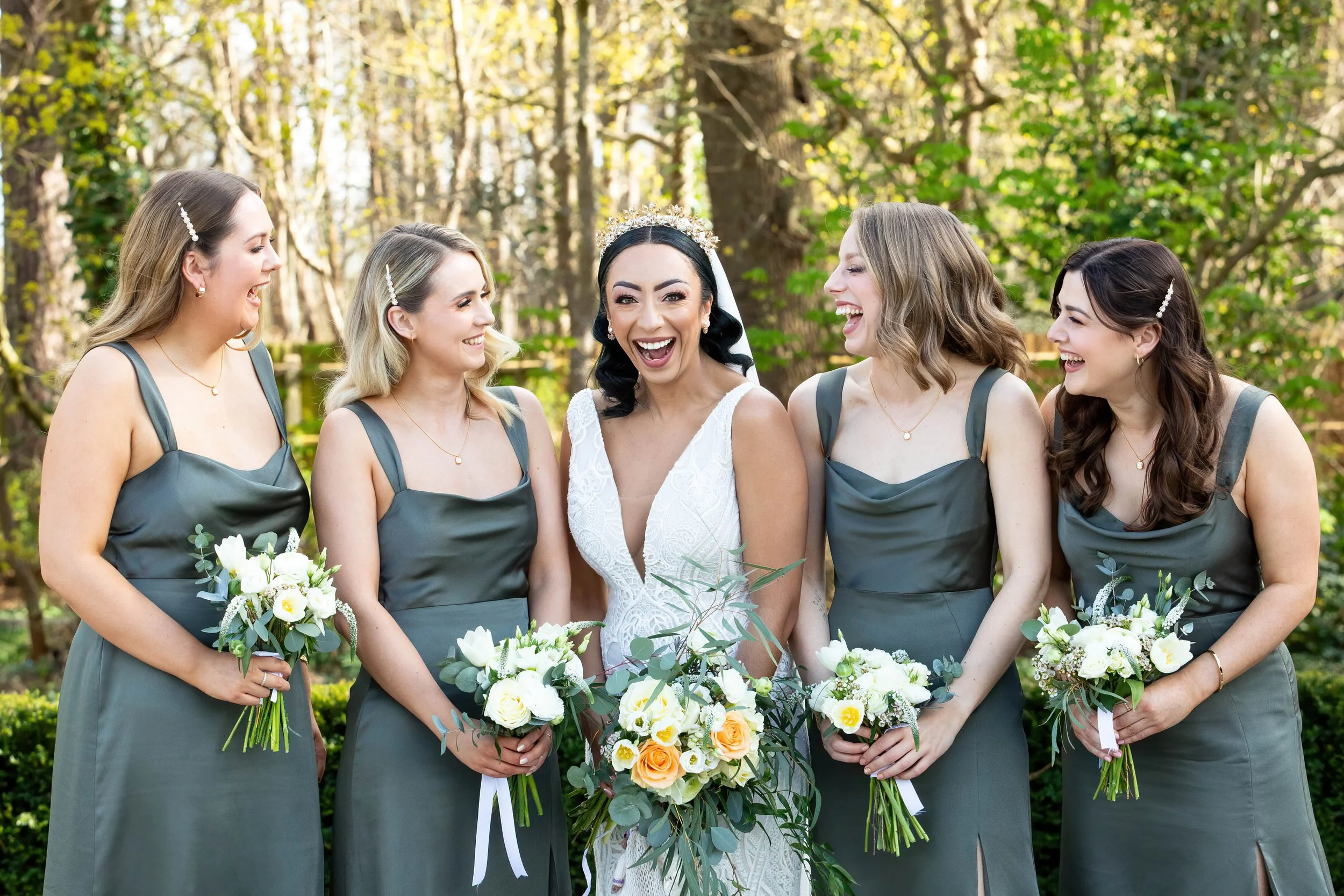 Bride in white wedding dress with tiara surrounded by four bridesmaids in matching grey dresses, holding bouquets of white and yellow flowers, outdoors in a wooded area.