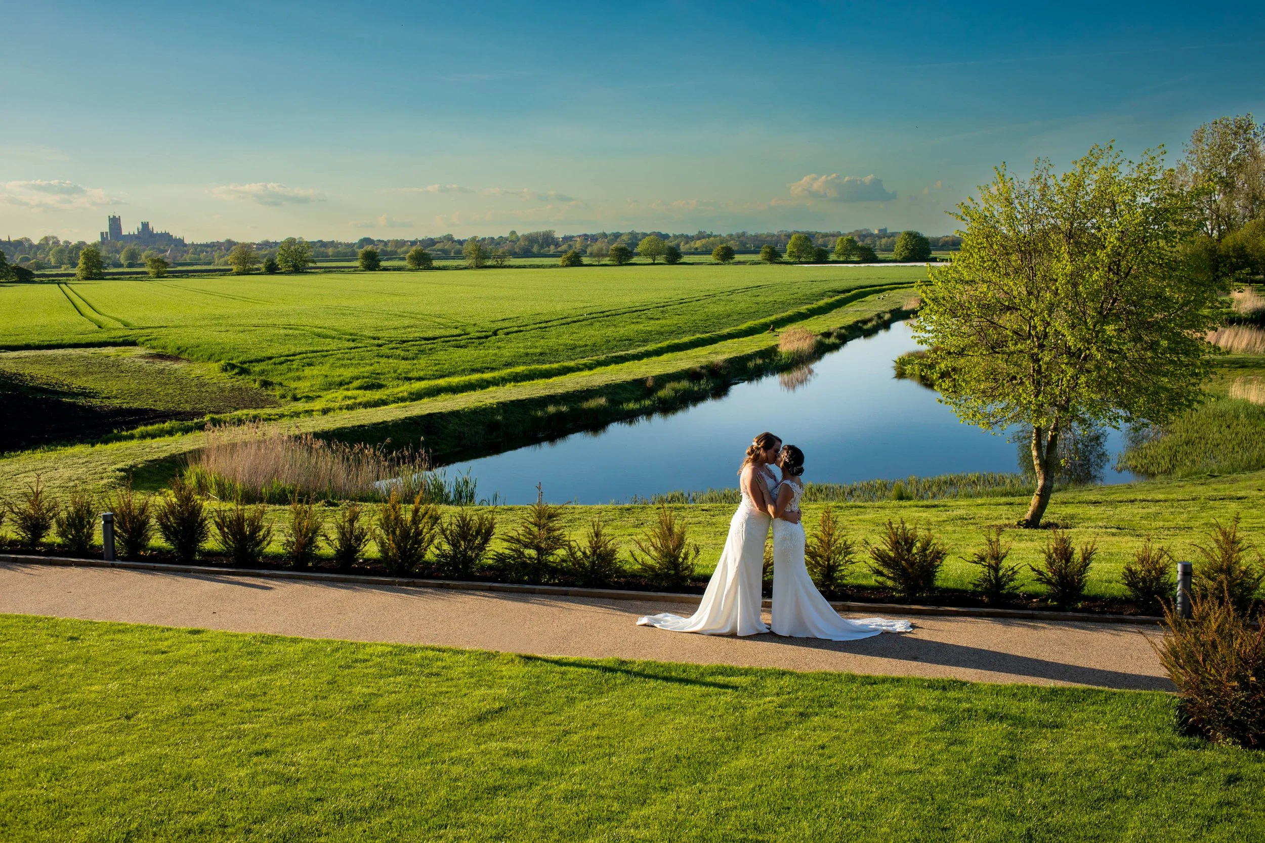 Two newly married brides in wedding dresses kissing in a park with green grass, trees, a pond, and a distant Ely Cathedral.