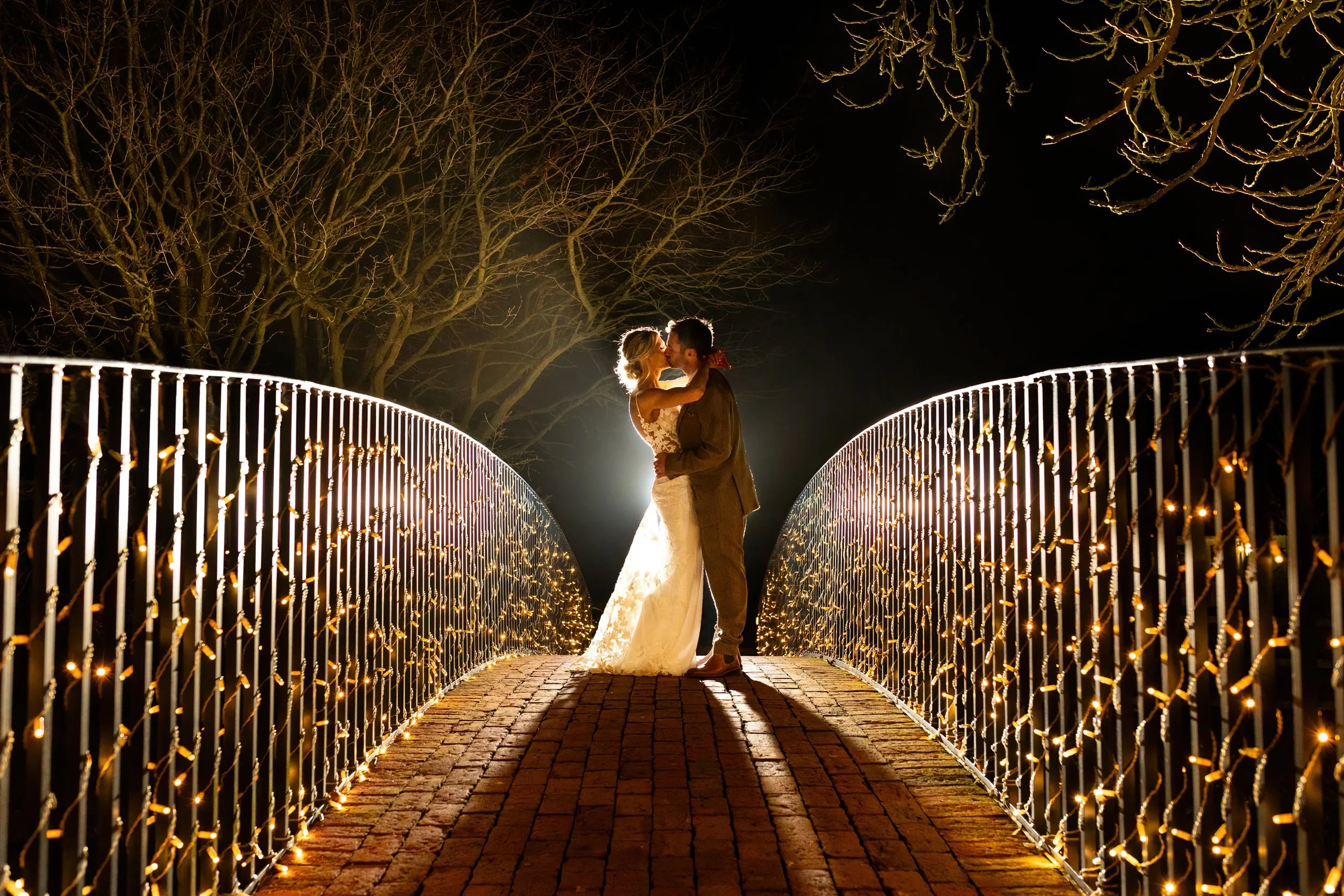 Bride and Groom sharing a kiss on a bridge at night at Bassmead Manor Barns, a romantic evening wedding portrait at the Cambridgeshire barn venue. 
