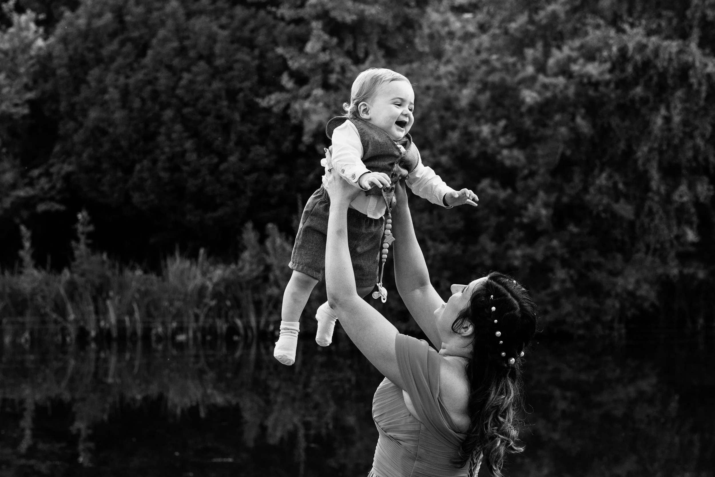 A woman lifting a smiling young child in the air outdoors near a body of water with trees in the background.