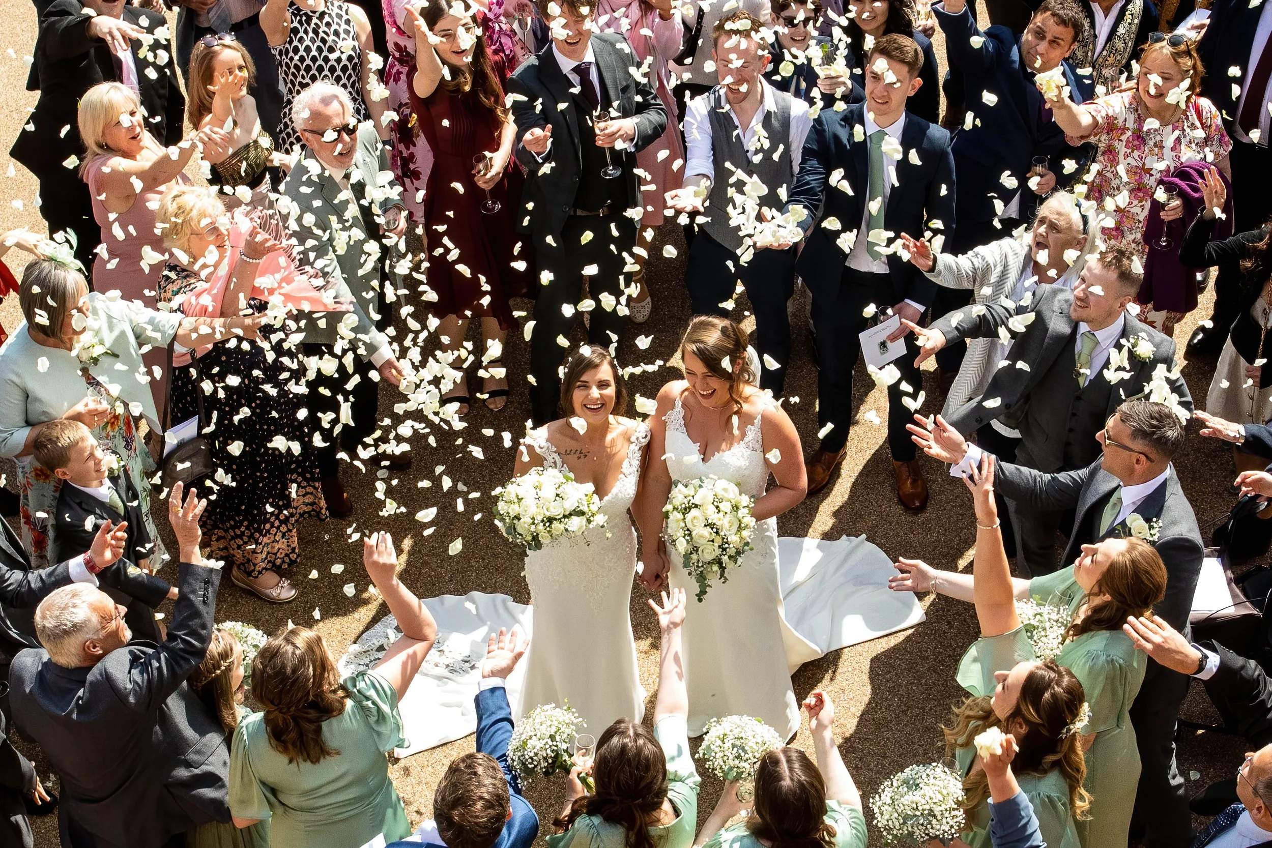 Two brides in white wedding dresses holding bouquets, standing together surrounded by guests throwing flower petals in celebration.