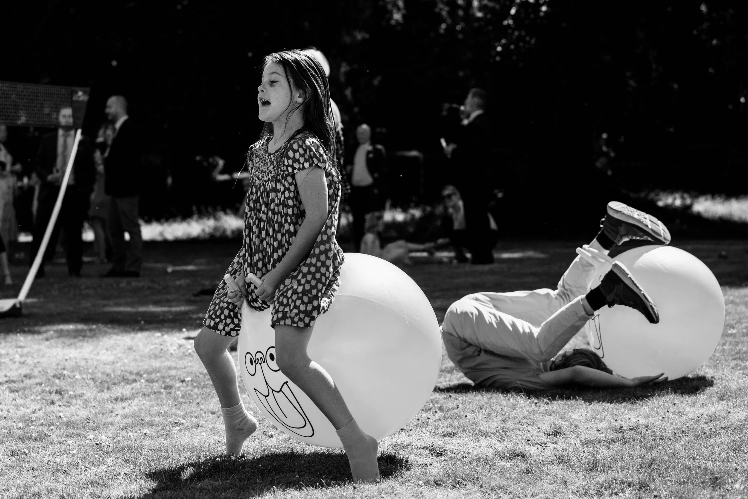 A girl in a polka dot dress bouncing on a large ball with a cartoon face, while a boy in white trousers is falling into another large ball on grass, and several adults are in the background.