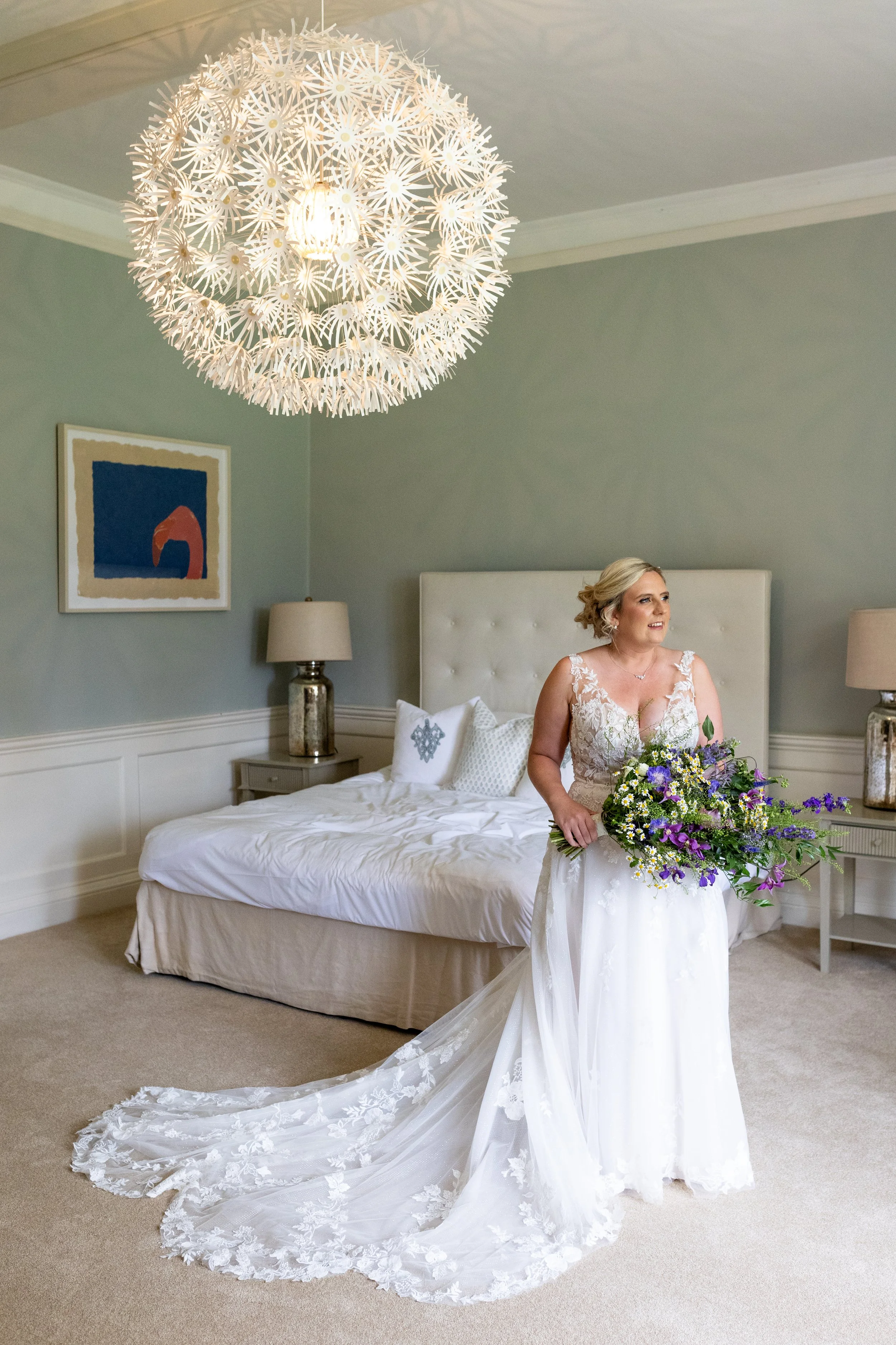 A bride in a wedding dress holding a bouquet of purple and white flowers, standing in a bedroom with a white bed, lamps, and artwork on the wall.