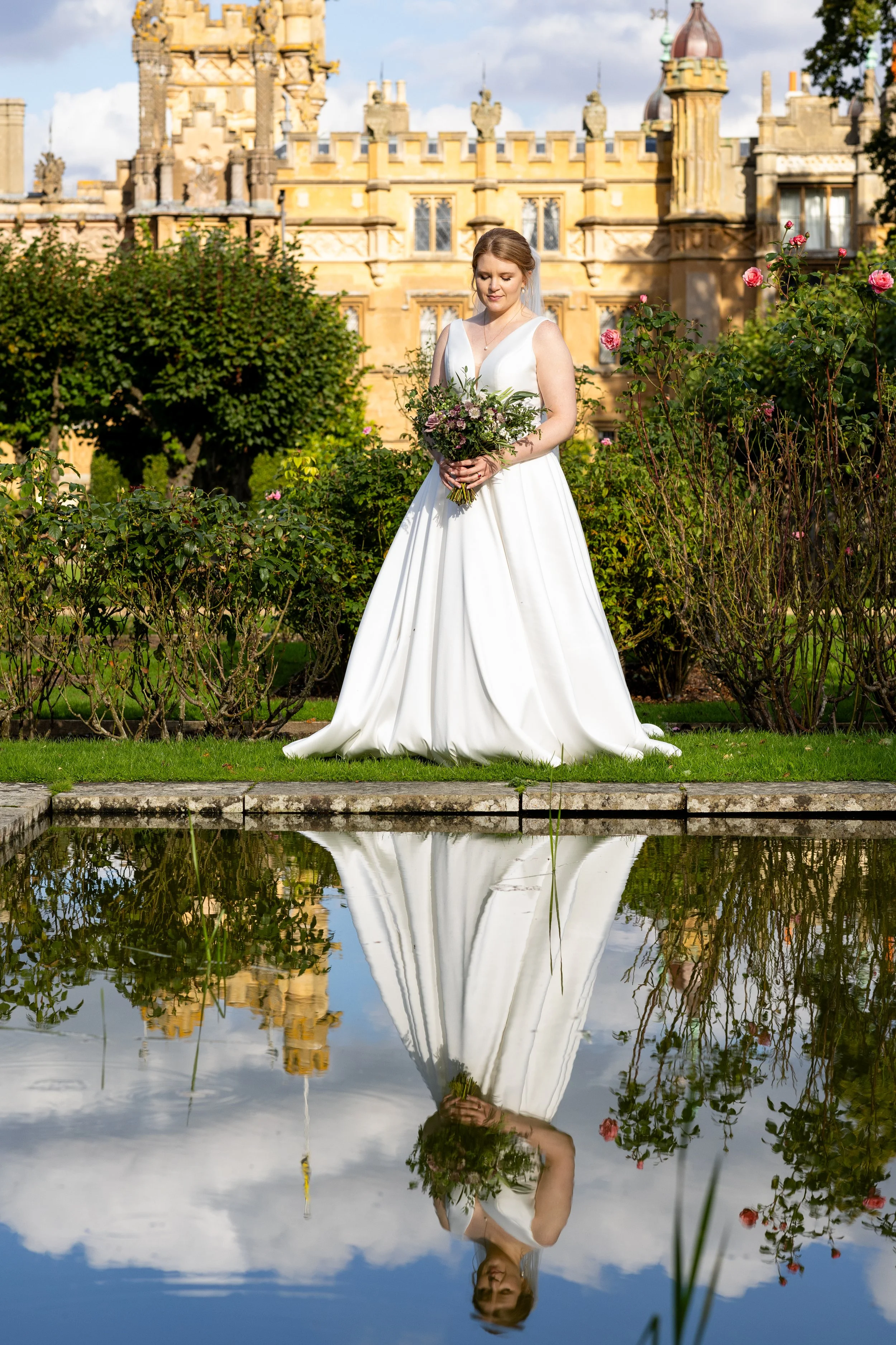 Elegant bridal portrait at Knebworth House Gardens, with the bride holding her bouquet beside the water and her reflection visible on the surface. 