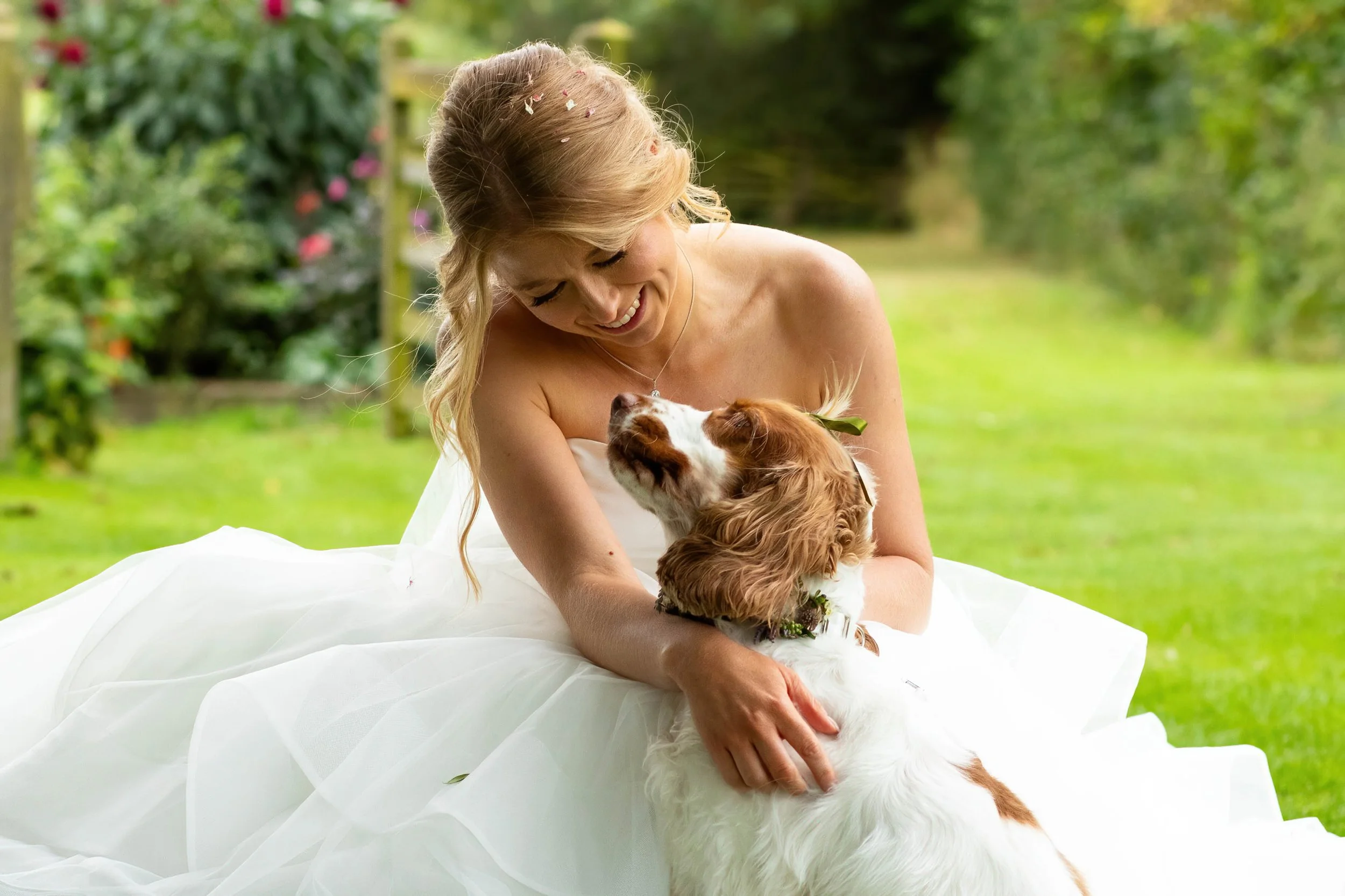 A bride dressed in a white wedding gown smiling and holding a dog in an outdoor garden setting.