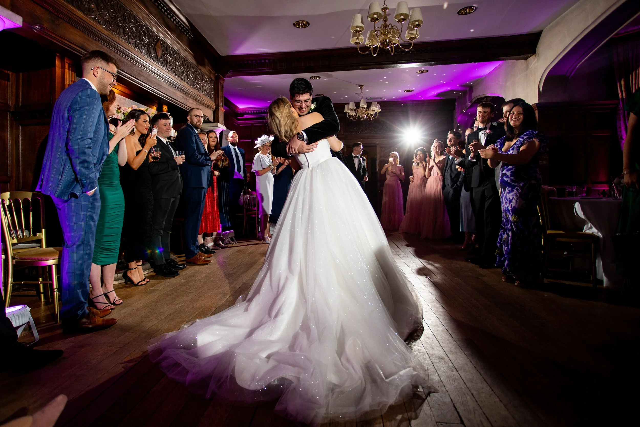 A bride and groom are dancing and hugging during their wedding reception. They are surrounded by party guests standing in a semi-circle, clapping and watching them. The room has elegant wooden architecture, purple lighting, and a chandelier hanging f