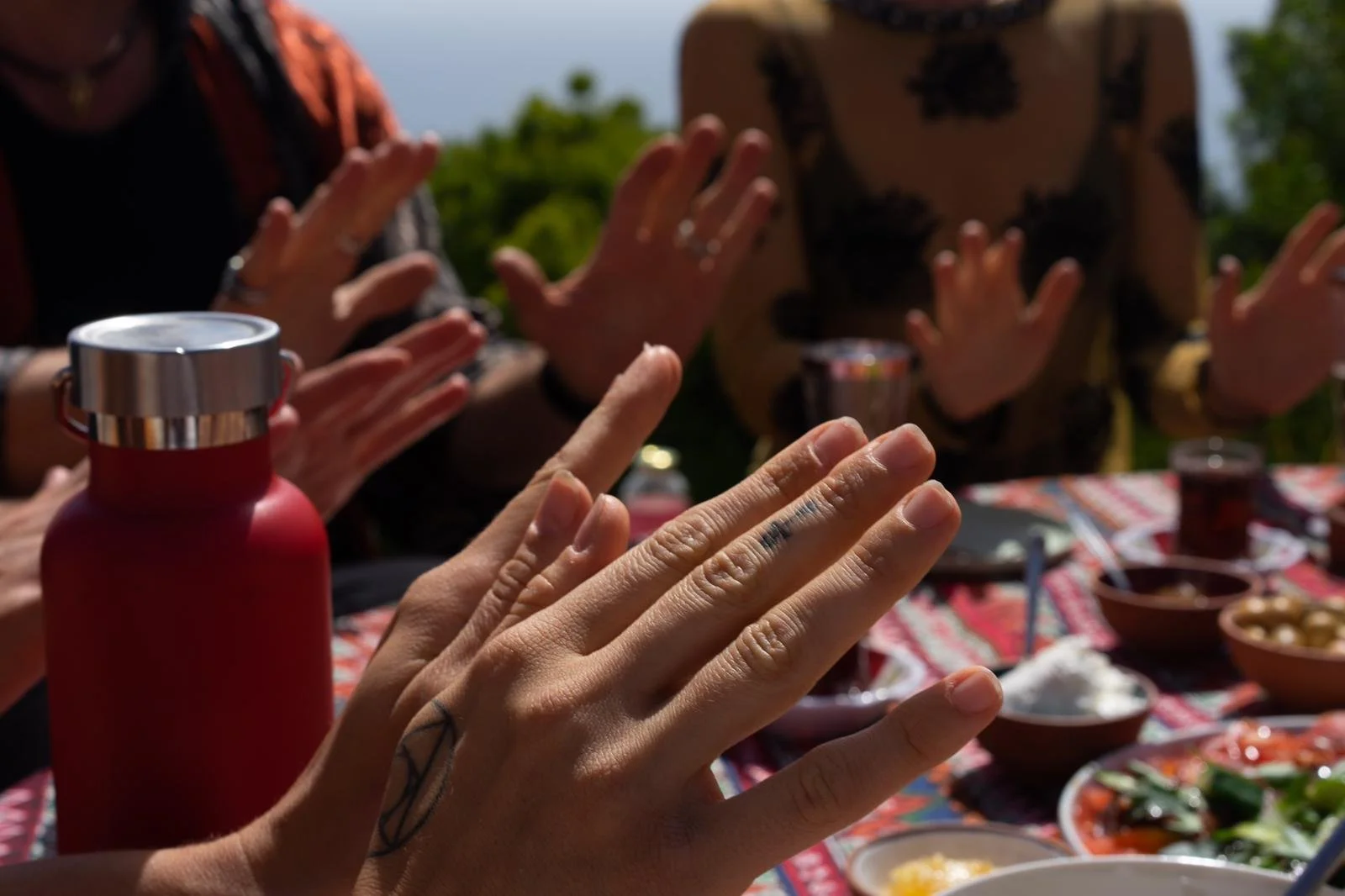 People with outstretched hands seated around a table with food, drinks, and bowls, outdoors on a sunny day.