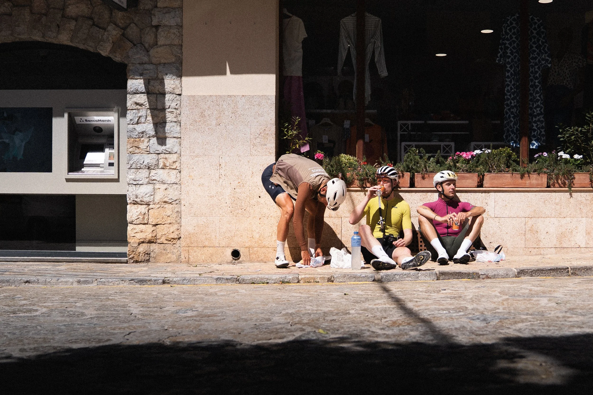 Three cyclists taking a supermarket stop on a long ride, eating and resting in the sun.