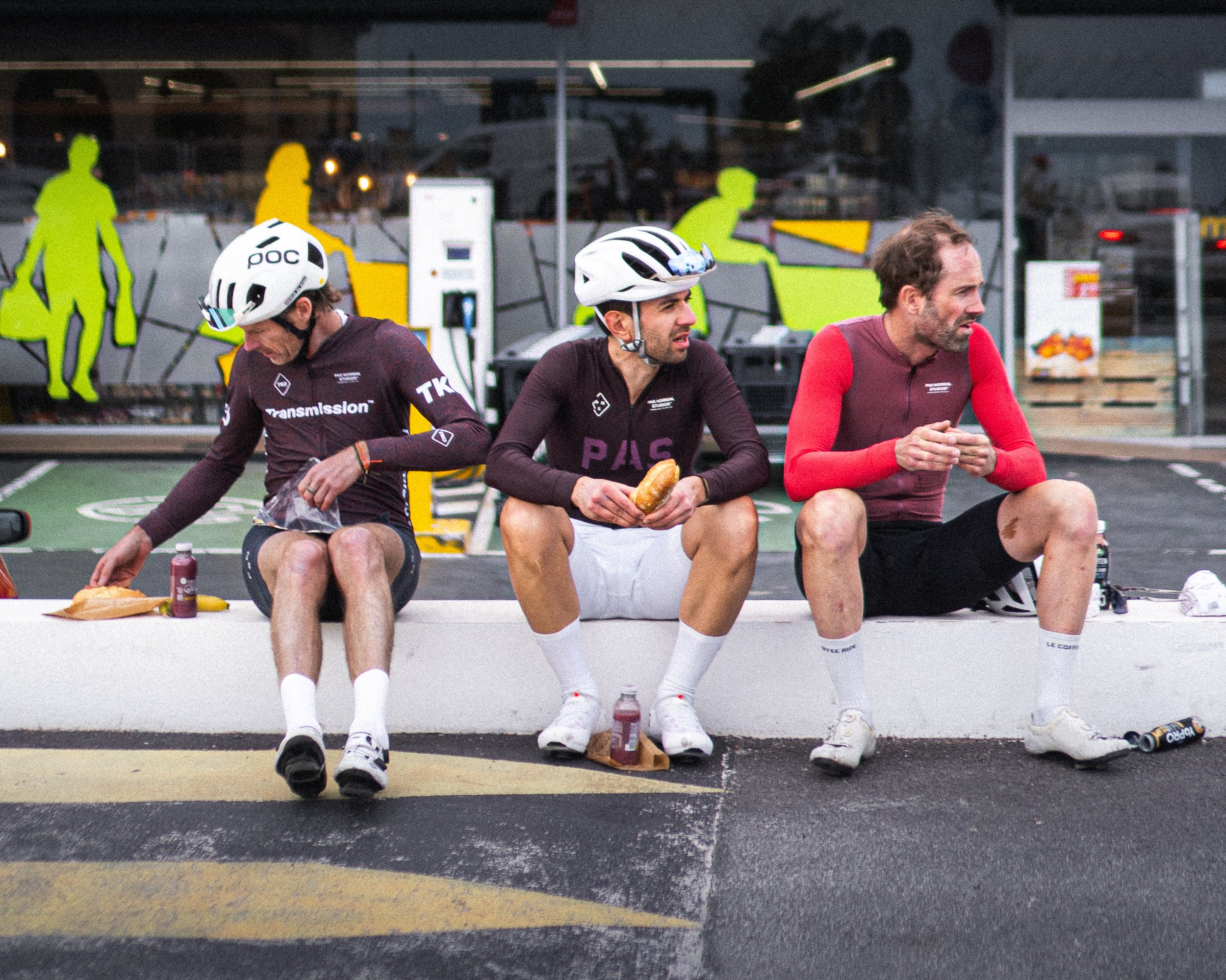 Three cyclists sitting on a kerb outside a supermarket during a long ride stop, eating and resting before rolling out again.