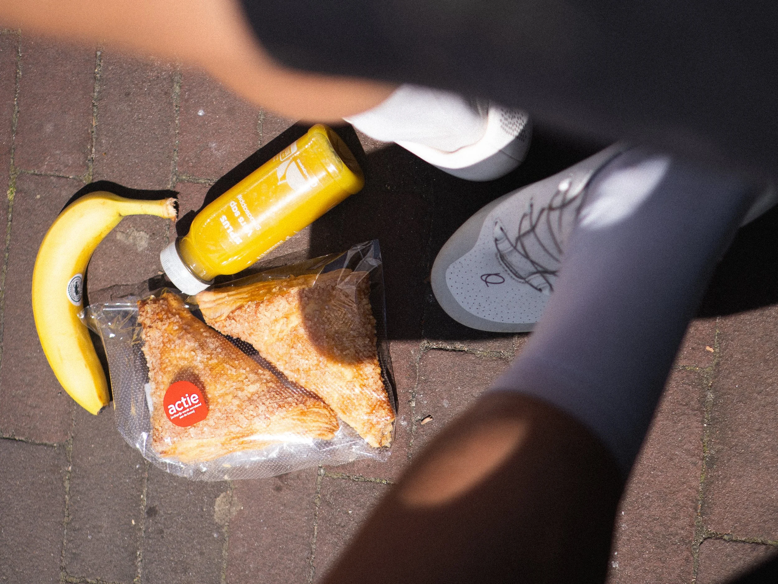 Cyclist sitting with supermarket stop purchases on the ground: a banana, a bottle of fresh orange juice and a Dutch pastry.
