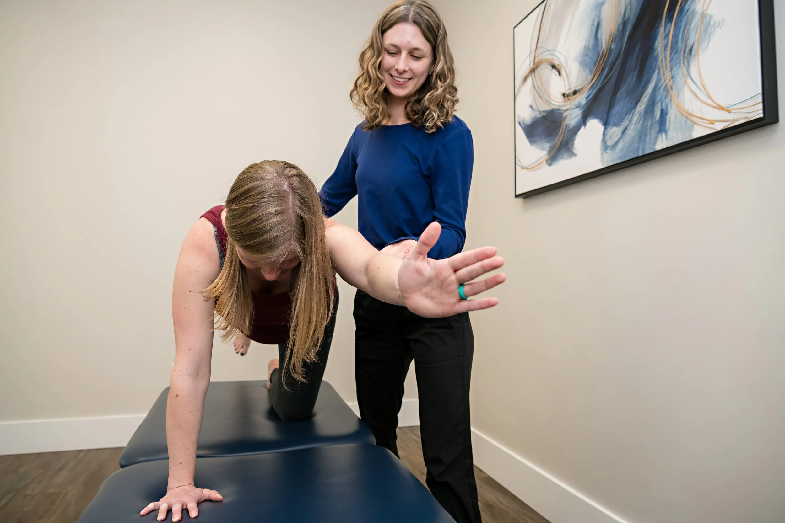 A physiotherapist is helping queue a client's plank pose on a treatment table in a clean medical office.