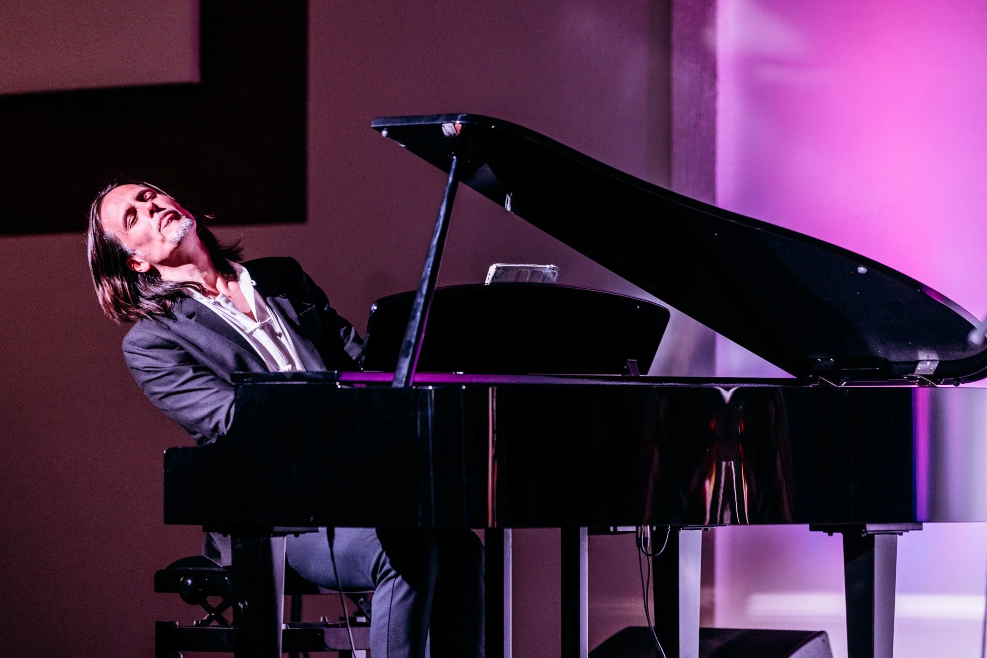 A man with long dark hair and a beard playing a grand piano, eyes closed, in a performance setting with pink and purple lighting.