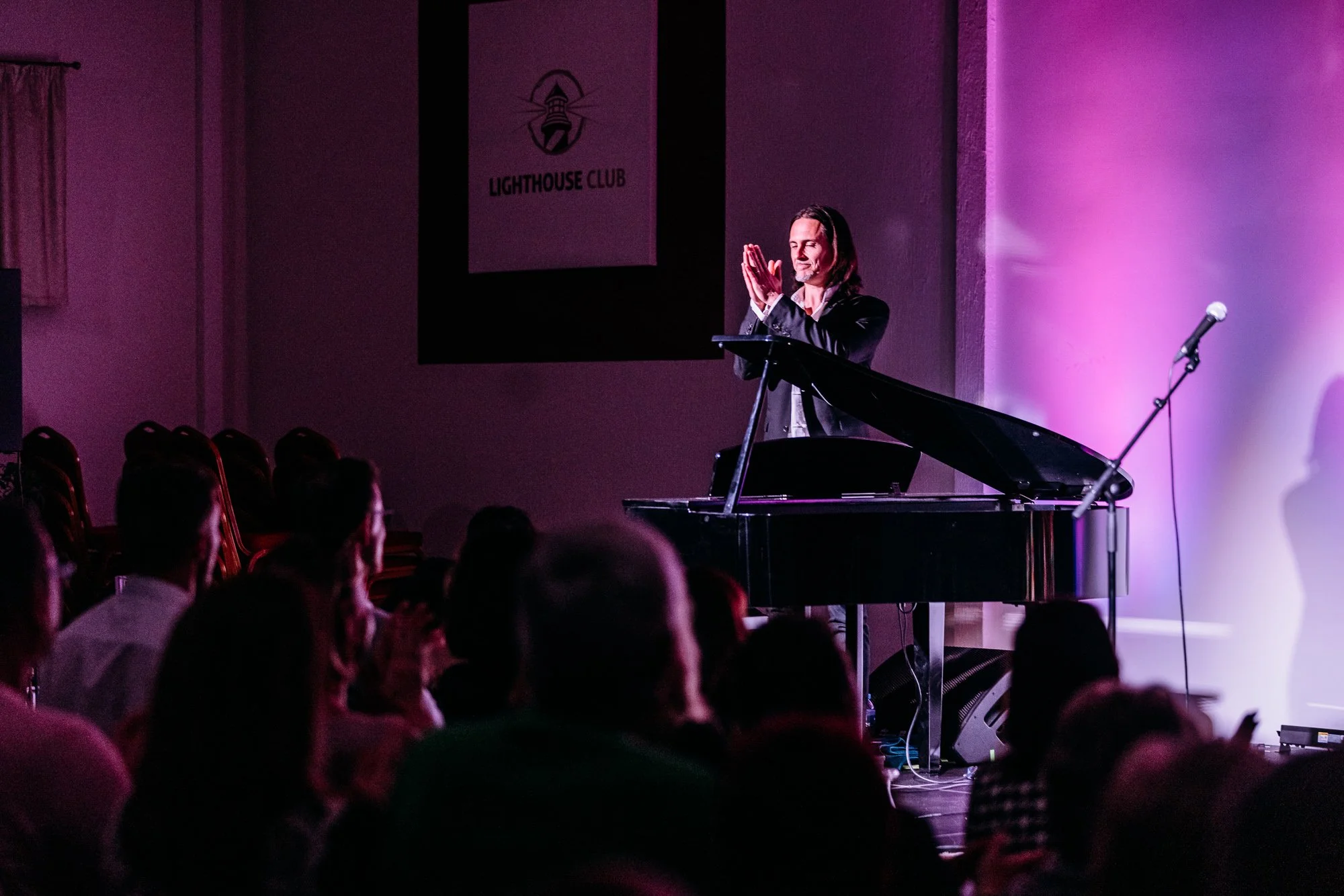 A performer standing behind a grand piano on a stage with purple lighting, gesturing with hands together in front of an audience at the Lighthouse Club.