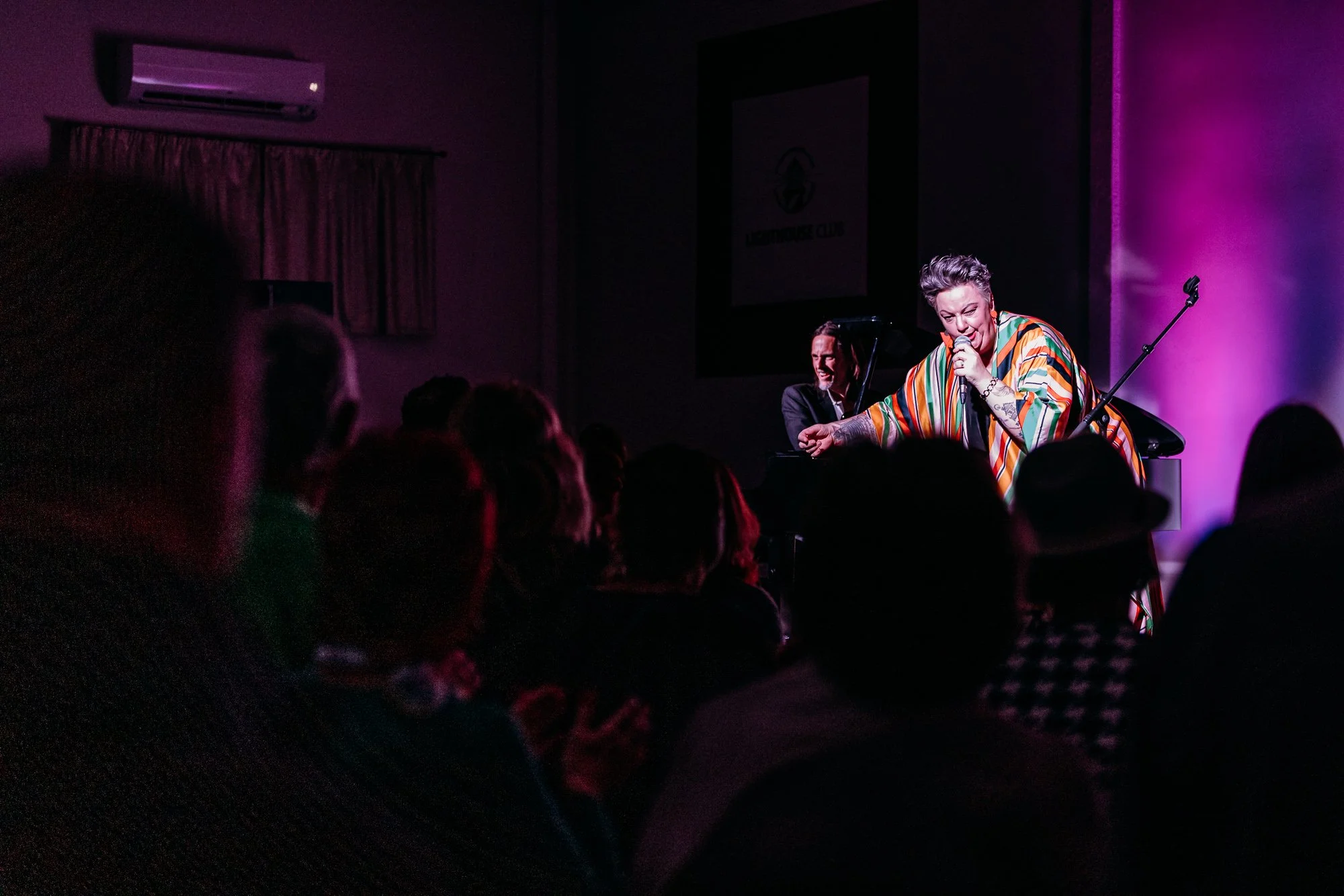 A singer with short gray hair and tattoos performs on stage, wearing a colorful striped dress, while a pianist in a dark suit plays in the background. The stage is lit with purple lighting, and an audience watches the performance.
