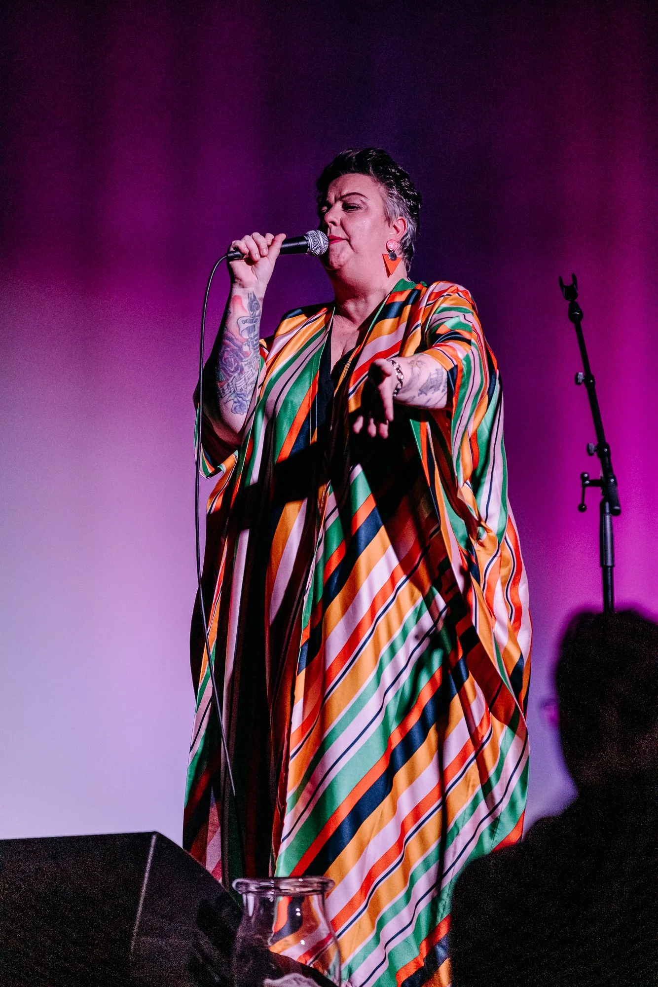 A person singing into a microphone on stage with a colorful, striped outfit against a purple background.