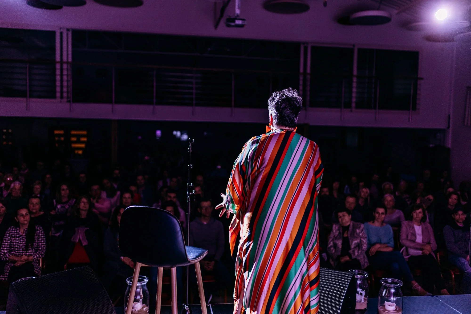 Performer on stage speaking to an audience in a dimly lit auditorium.