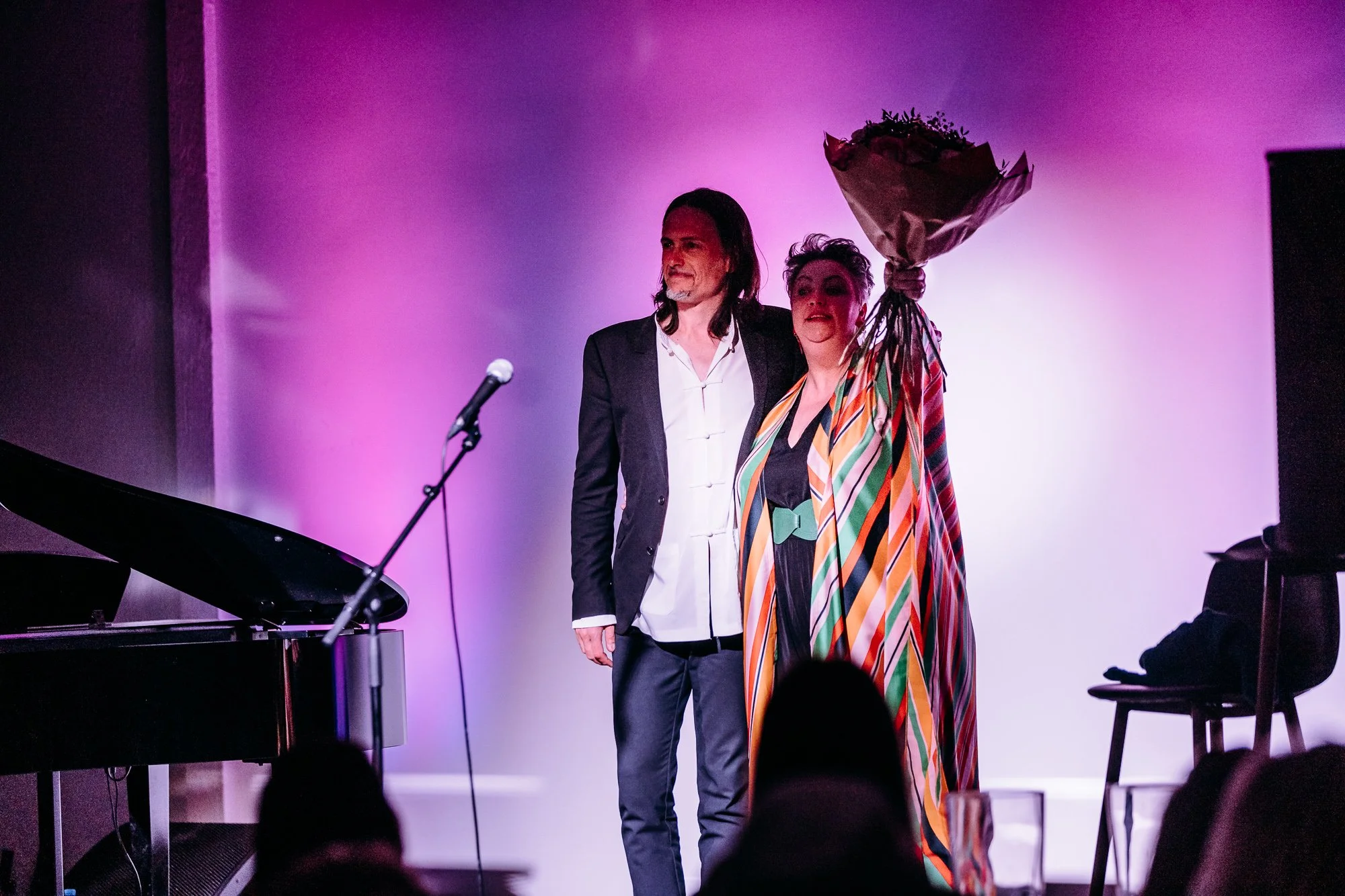 A man with long hair and beard wearing a dark blazer and white shirt standing next to a woman with short hair in a colorful striped dress holding a bouquet of flowers on a stage with purple lighting, a piano, and a microphone.