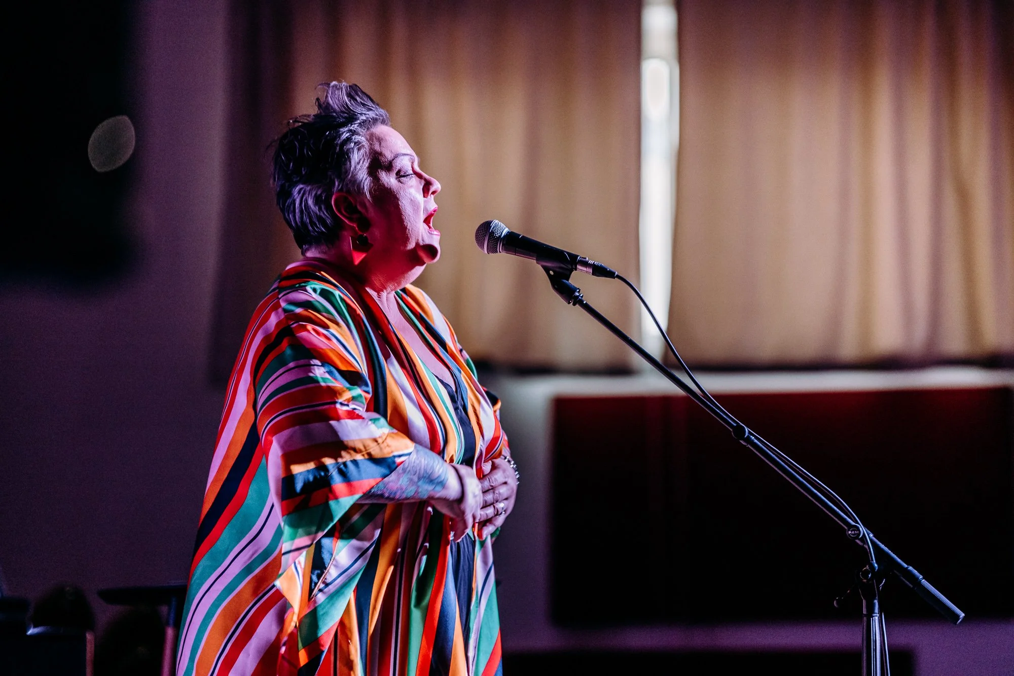 A woman with short gray hair singing into a microphone, wearing a colorful, striped dress, standing in front of closed beige curtains.