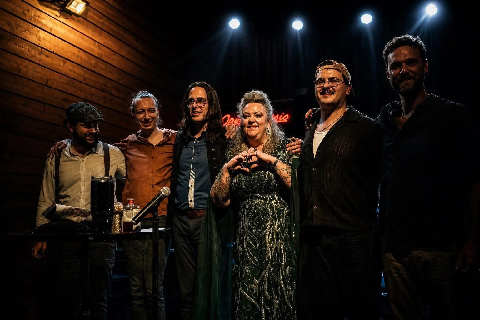 Group of six people standing on stage with dark background and lighting, some with arms around each other's shoulders, smiling, in a bar or music venue setting.