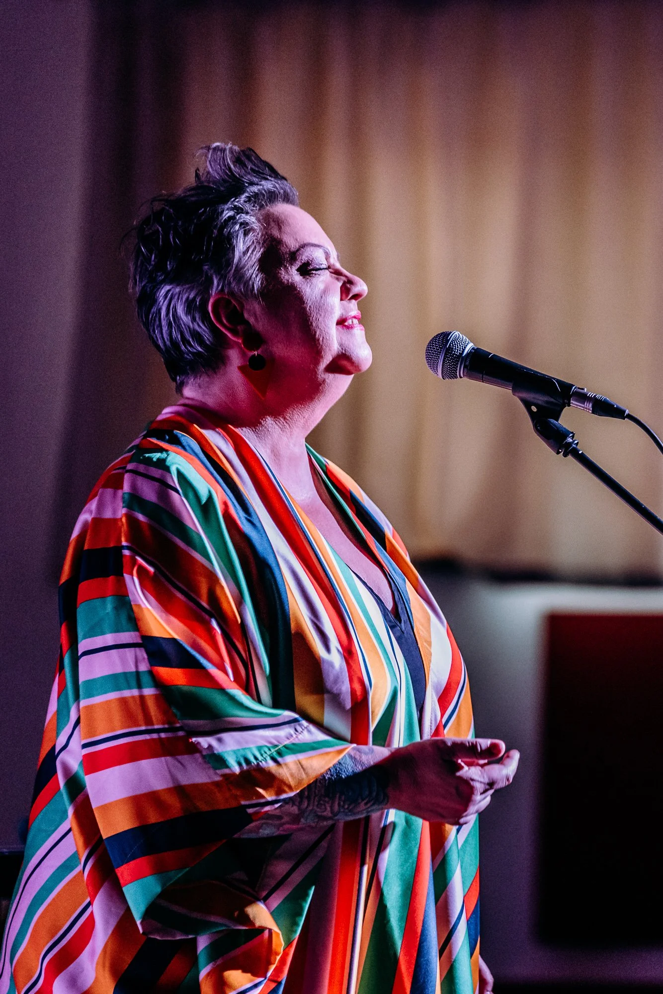 A woman with short, gray hair singing into a microphone on stage, wearing a colorful, striped blouse with shades of red, green, orange, and black.