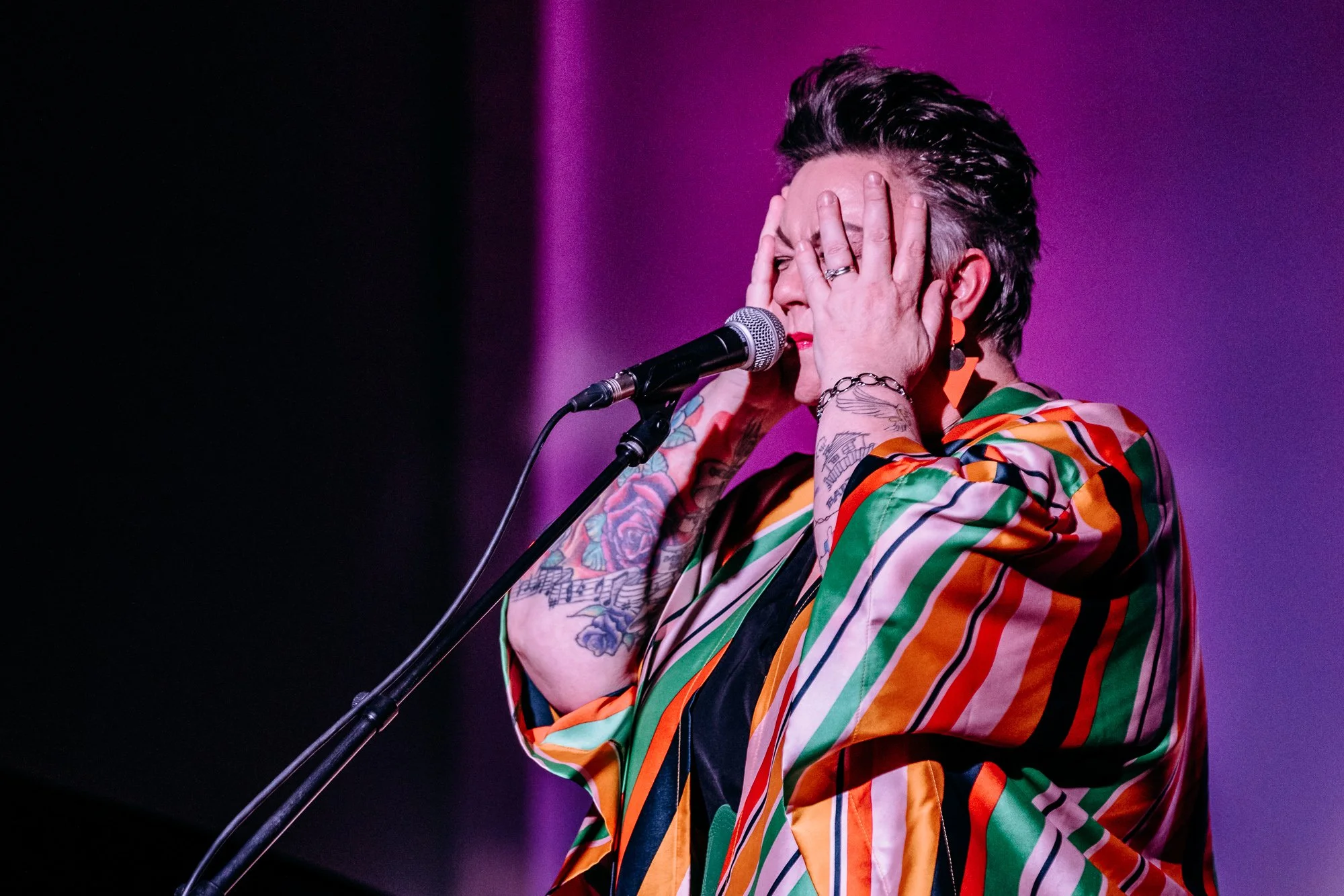 A woman with short, styled hair singing into a microphone with her hands on her face, wearing a colorful striped jacket and showing tattoos on her arms.